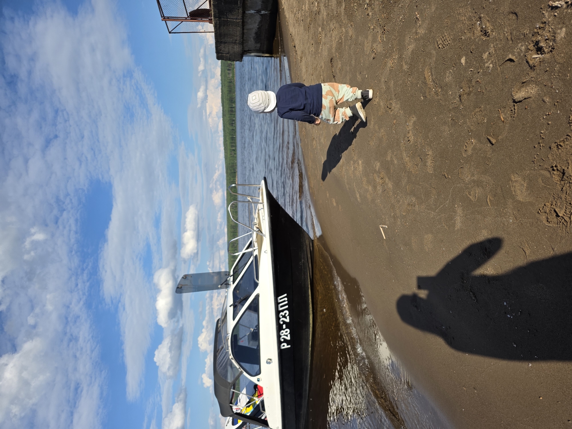 A child wearing a white cap, a dark blue long-sleeved shirt, and patterned pants stands on a sandy shore facing a body of water. A black and white motorboat with the registration number "P28-23 ПЛ" is partially beached on the left side of the image, with its bow in the shallow water. The shoreline is made of dark sand with some debris. In the background, across the water, a dense green forest line is visible under a blue sky with scattered white clouds. To the right, a concrete structure with a metal fence extends into the water. The shadow of a person holding a phone is cast on the sand in the foreground. The location appears to be a waterfront area in Chaikovsky, Russia, likely a pier or a small harbor.
