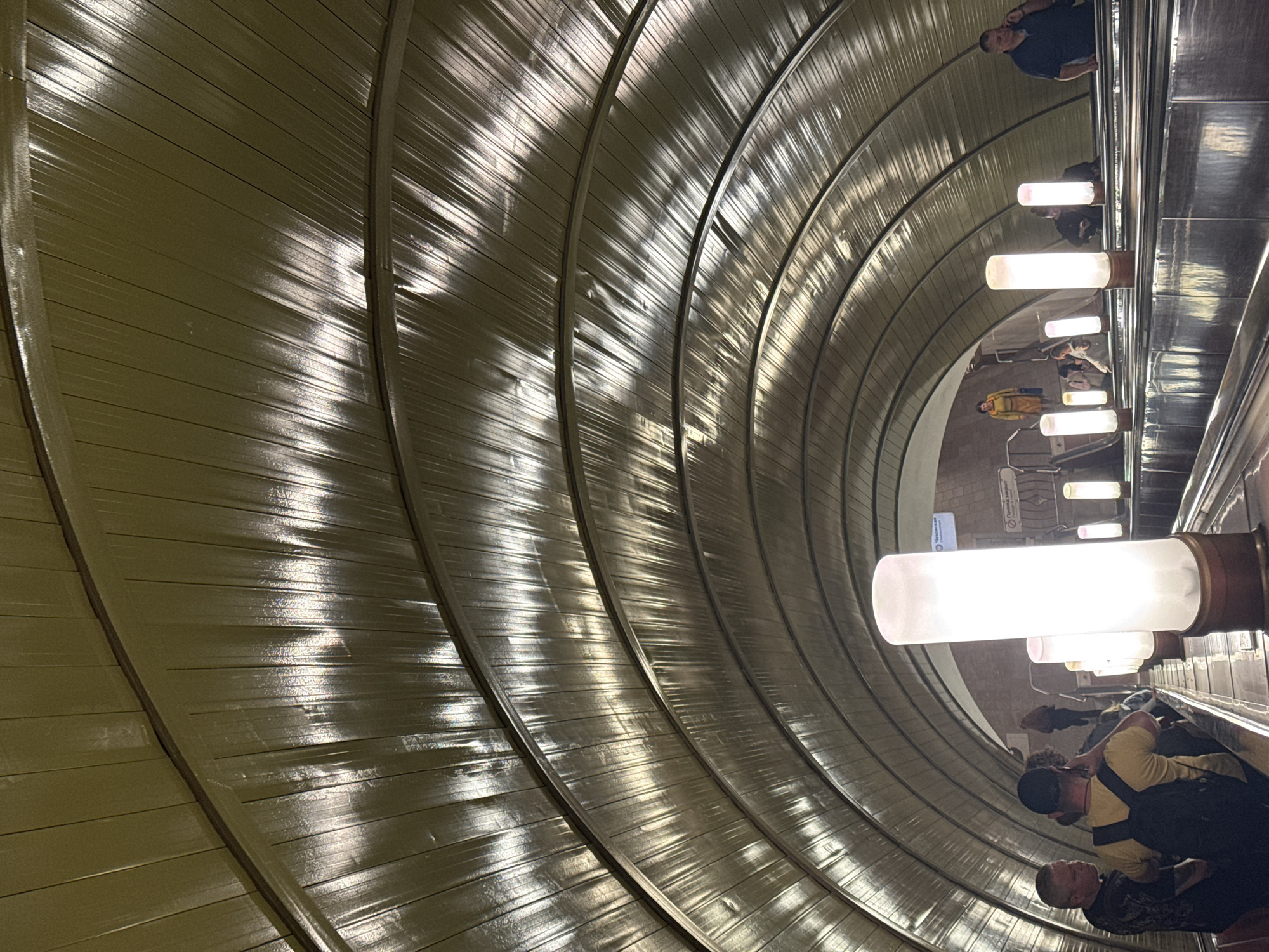 The image captures a deep, curving escalator tunnel, likely within a Moscow Metro station, viewed from a high vantage point looking downwards. The most striking feature is the dramatic, ribbed ceiling or wall structure that arcs overhead, displaying a greenish-brown, metallic, or laminated surface. This surface intensely reflects the artificial light, creating luminous streaks and patterns along its numerous parallel ridges, emphasizing the tunnel's curved depth.
A series of bright, cylindrical white lights are mounted horizontally along the right side of the tunnel, illuminating the scene. Several individuals are visible on the escalator, appearing to descend. Among them, a man in a dark blue shirt is seen towards the upper right. Further down, another person is wearing a yellow top, and closer to the bottom, a man in a black cap and yellow t-shirt is visible with a black backpack. The people seem to be commuters or travelers.
On the left side of the tunnel wall, in the mid-ground, there are signs with Cyrillic text. One clearly legible sign includes the word "Черкасская" (Cherkasskaya), indicative of a station or destination, and another sign below it reads "Проход закрыт" (Passage closed). The architecture, the depth of the escalator, and the visible text confirm the location as part of the Moscow Metro system, known for its distinctive and often ornate underground stations. The scene is well-lit, highlighting the unique architectural details and the movement of people within the grand, functional space.