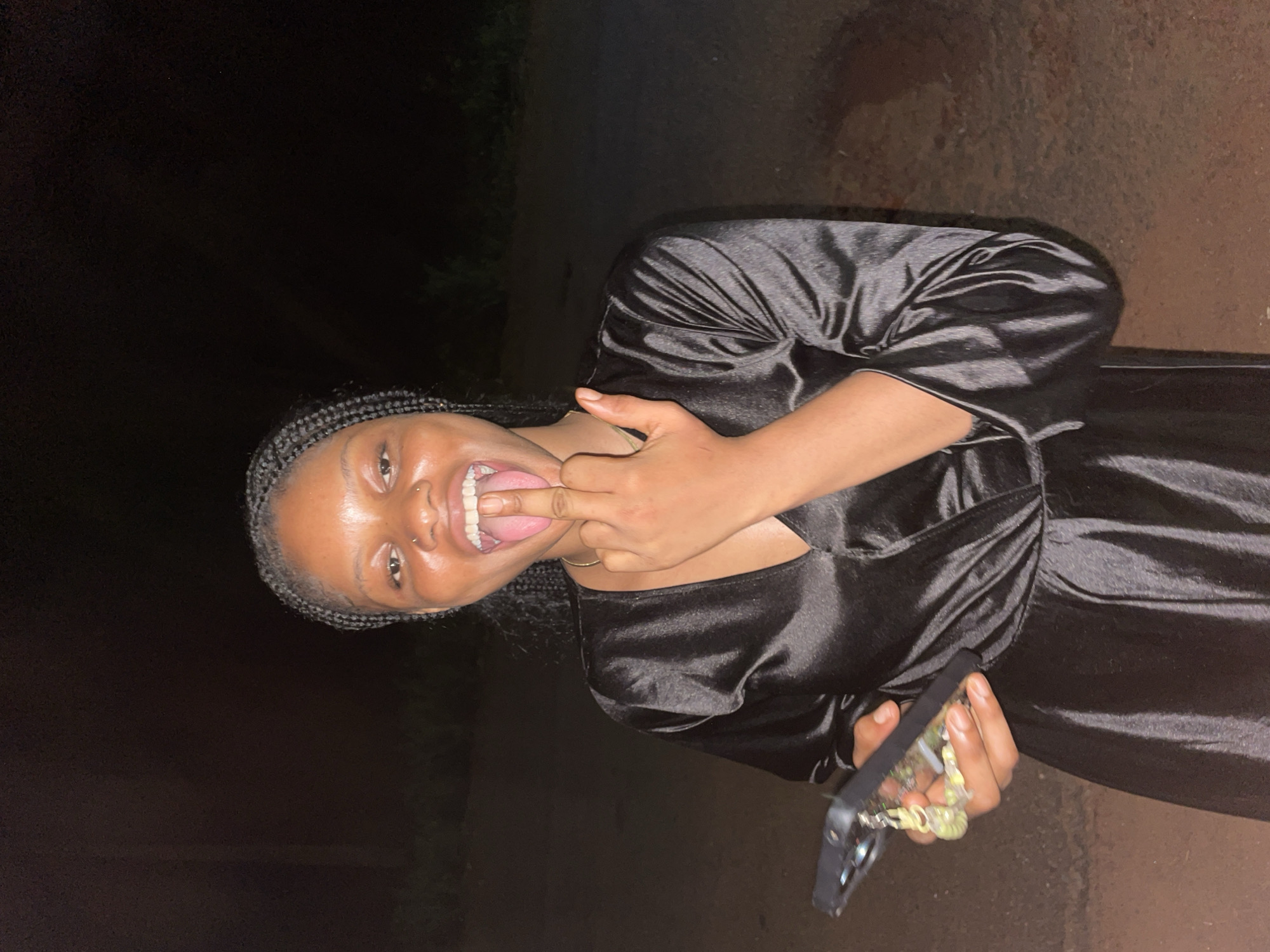 A young Black woman is captured in a nighttime portrait, likely in Ogbe, Nigeria. She stands facing the camera, wearing a shiny black, long-sleeved dress, possibly made of satin or silk. Her hair is styled in braids. She displays a wide, joyful smile, her tongue extended outwards, while her right hand is raised with a finger touching her tongue in a playful gesture. In her left hand, she holds a smartphone encased in a clear cover, adorned with a decorative, possibly beaded, charm or strap. She has a small piercing on her nose. The background is very dark and indistinct, suggesting an outdoor setting in low light conditions, with hints of a dark, uneven surface on the ground behind her. No discernible text is visible in the image.