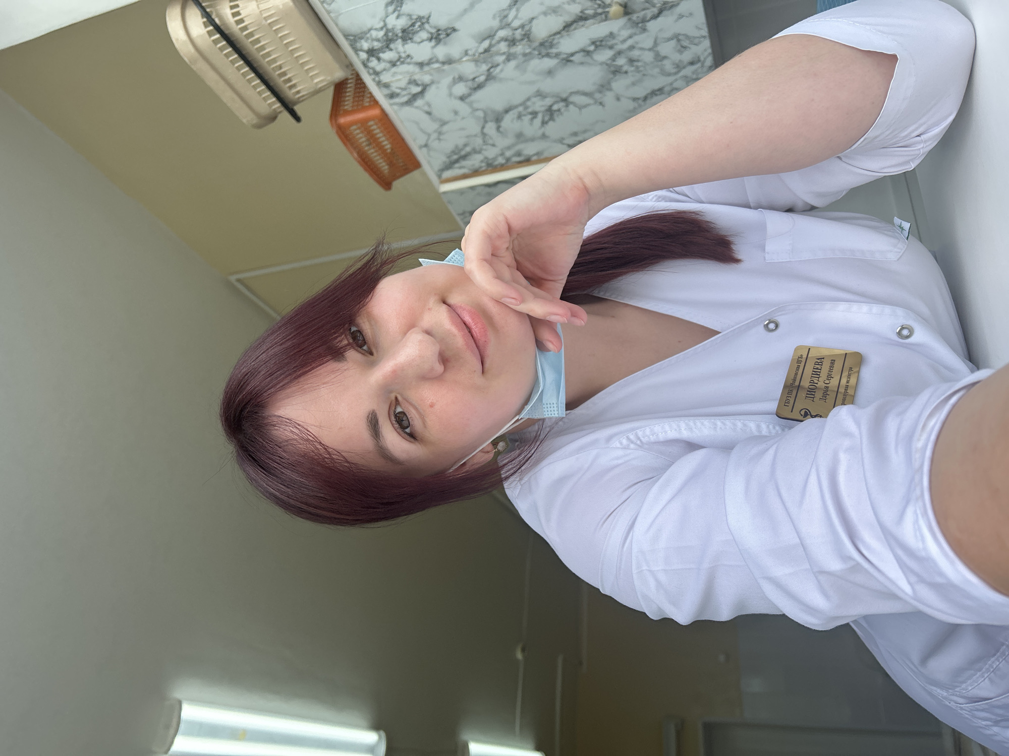 This is a selfie taken by a young woman wearing a white medical coat. She has long, dark red hair and is looking towards the camera with a neutral expression. She is wearing a light blue surgical mask that is partially pulled down below her nose. Her right hand is resting on her cheek. A name tag is attached to her lab coat, which appears to be for a medical professional. The text on the name tag is in Russian and reads: "ГБУЗ «Чижовская районная больница»", "ДИОРДИЕВА Дарья Сергеевна", and "помощник врача, медсестра". The setting appears to be indoors, possibly a medical facility, with tiled walls in the background and fluorescent lighting. There are also some plastic baskets visible on a shelf in the background. The lighting suggests it could be daytime.
