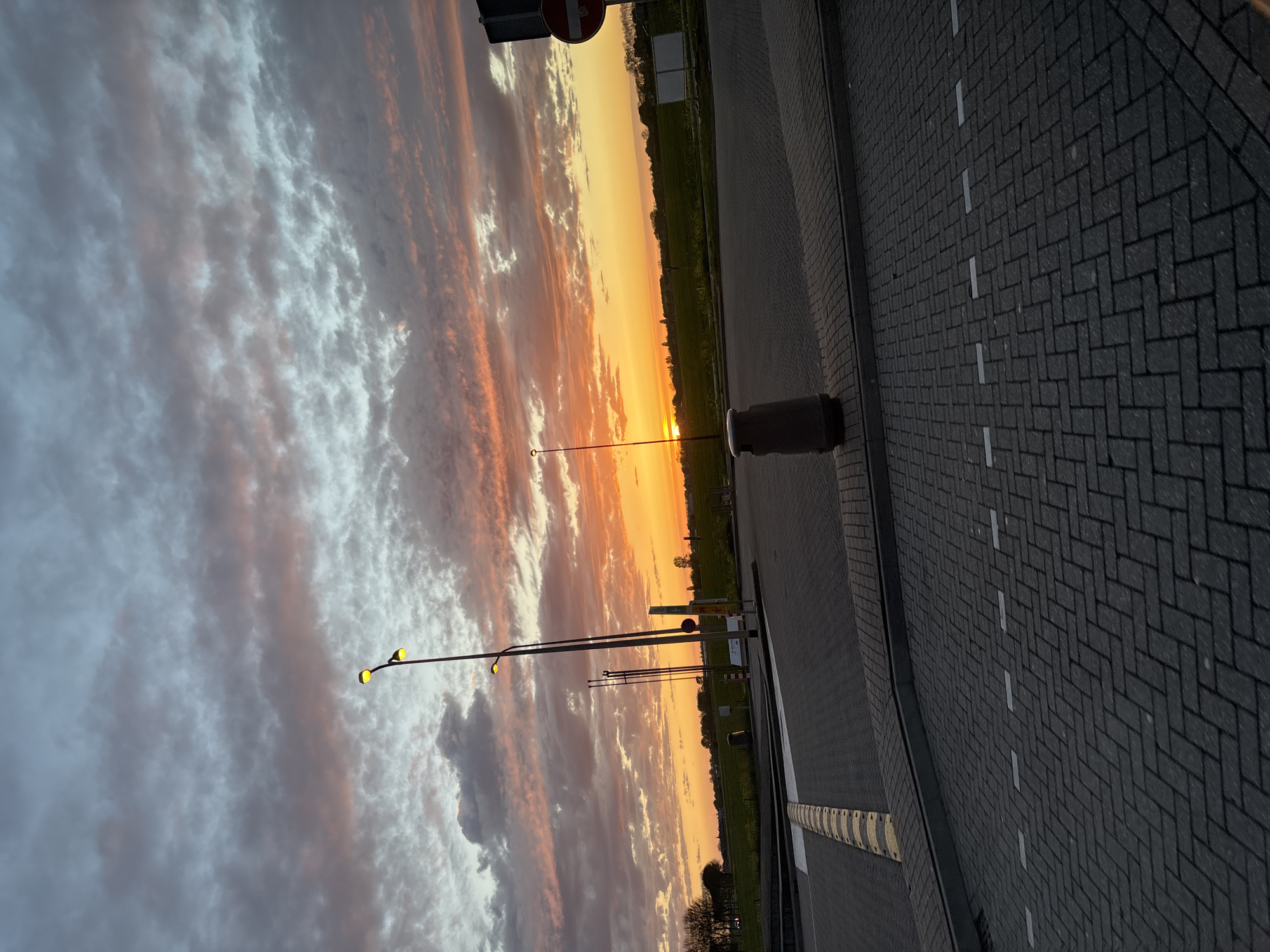 The image depicts a sunset scene in Bruchem, Netherlands. The sky is filled with colorful clouds in shades of orange, pink, and gray. Streetlights are visible on the left side of the image, with their yellow lights illuminated. A paved area with brick-like patterns occupies the right side of the image, with white lines marking the edge. A cylindrical object, possibly a bollard or trash receptacle, is located on the edge of the pavement. A strip of green land is visible between the sky and the paved area, with some vegetation and trees. A road sign is seen in the upper left corner. The scene is captured at dusk or dawn.