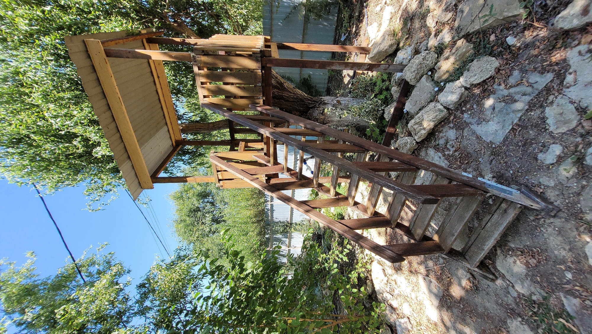 An outdoor shot shows a wooden treehouse with a pitched metal roof, supported by a large tree and two wooden posts. A set of wooden stairs with a handrail leads up to the treehouse platform. The treehouse and stairs are made of weathered brown wood. The area around the treehouse is partially paved with large flat stones and covered with dirt, leaves, and small rocks. The background features a metal fence, dense green foliage, and a bright blue sky. The scene appears to be a yard in Gostagayevskaya, Russia, on a sunny day. There are no people or animals visible.