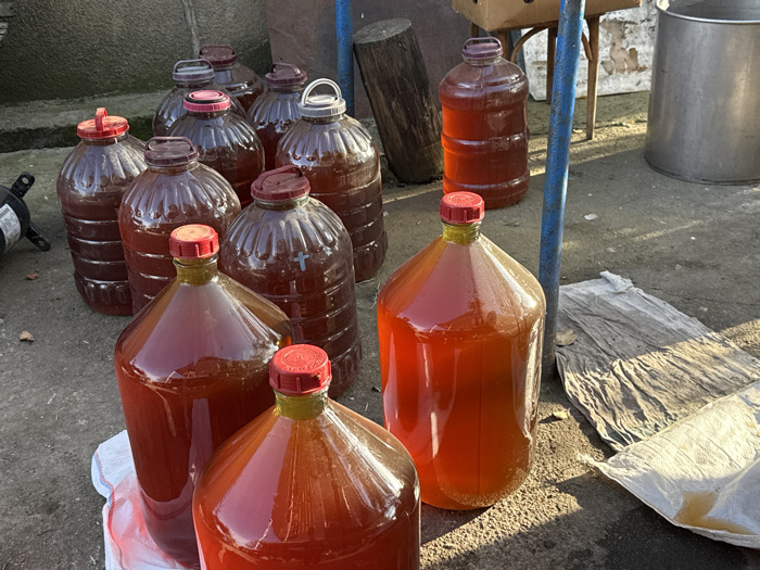 The scene is set outdoors in Bakurtsikhe, Georgia, displaying approximately fifteen large plastic carboys filled with a translucent, reddish-brown liquid. The carboys vary in size, with most appearing to be 5-gallon capacity or larger. They are equipped with screw caps, predominantly red or dark brown, though one cap features a white handle, and another carboy bears an embossed cross symbol on its side.
These containers are arranged on an uneven, light grey concrete or compacted dirt surface, illuminated by direct sunlight that casts distinct shadows. In the background, to the left, is a textured grey wall structure. To the right, various objects are visible, including a blue metal pole, several wooden logs, a cardboard box resting on a wooden stool, and a large silver-colored metal container. A crumpled, stained white or light grey tarpaulin or sack lies on the ground in the mid-right. No individuals are present in the image.