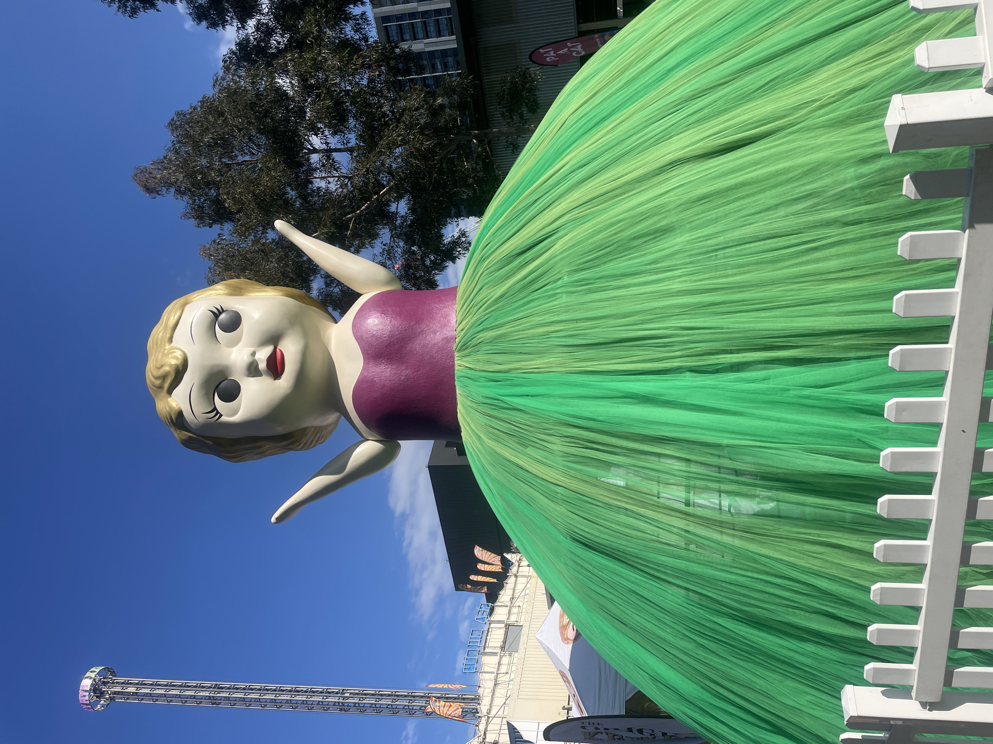 In Bowen Hills, Australia, a large, stylized doll sculpture stands outdoors, dominating the frame with its vibrant green tulle skirt. The doll has a rounded head with blonde hair, wide eyes with black pupils and white highlights, and red lips. It wears a purple bodice. The skirt, made of translucent green fabric, is voluminous and extends outwards, obscuring the lower part of the sculpture and a white picket fence in the foreground. Behind the doll, a tall, silver amusement park ride with a circular structure at the top is visible against a clear blue sky. Trees with dense foliage and a modern building with windows are also present in the background, suggesting an urban or entertainment district setting. The overall impression is of a whimsical, eye-catching installation in a public space.