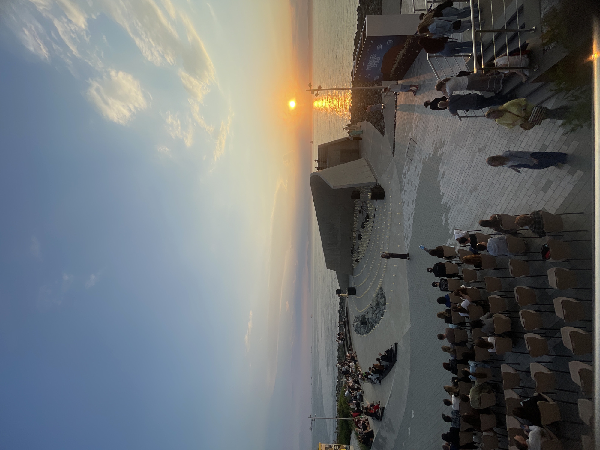 The image depicts an outdoor event with a sunset over a body of water. The scene is set in a modern open-air venue with tiered seating arranged in a semi-circle. Many people are seated, facing a stage or performance area. Another group of people are gathered on a paved walkway overlooking the water, some standing and some on steps.
The time of day is evident from the low sun casting a warm, golden glow across the sky and water, indicating sunset. The sky is partly cloudy, with wispy clouds tinged yellow by the setting sun. The water appears calm, with a reflection of the sun shimmering on its surface. Distant landmasses can be seen on the horizon.
A speaker or performer is visible on the stage area, and a few individuals are standing around the performance space, some appearing to be staff or organizers. The overall atmosphere seems to be one of attentiveness and calm observation as the audience watches the event unfold against the backdrop of the sunset. The location appears to be a waterfront promenade or amphitheater, possibly a public gathering space in Vladivostok, Russia, given the context.
The paved ground is decorated with numerous small, lit candles arranged in circular patterns. There is a large structure on the right side of the frame, possibly part of the venue's infrastructure or a display. Some text is visible on a blue banner on this structure, but it is too blurry to read. The general mood is peaceful and serene, enhanced by the beautiful natural light.