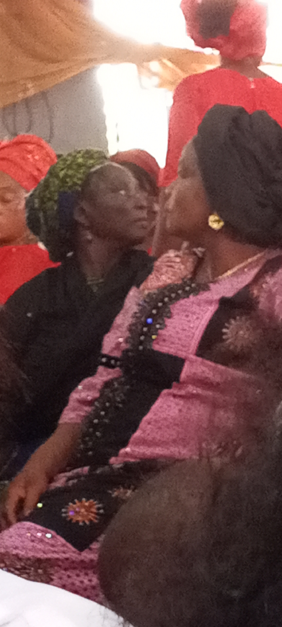 The image shows a group of people indoors. The woman in the foreground is wearing a pink patterned dress with black trim and a black head covering, and she has gold earrings. Another woman behind her is wearing a black outfit and a green and blue head wrap. In the background, there are other people wearing red clothing and head coverings. The location is Sabon Tasha, Nigeria.