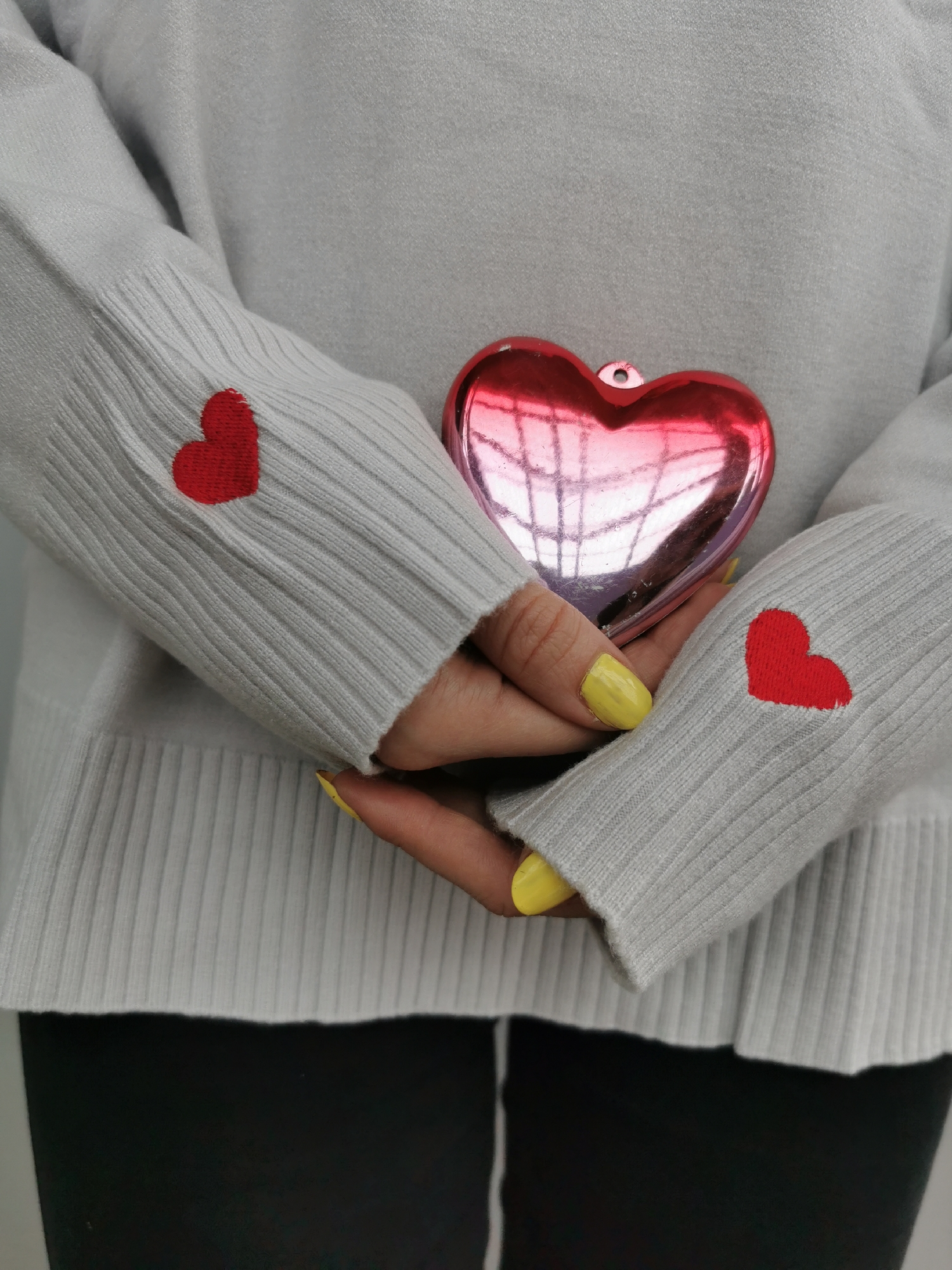 A person wearing a light grey sweater with red hearts on the sleeves is holding a shiny red heart-shaped object. The person has yellow nail polish. The background appears to be a solid, neutral color. The location is unknown.