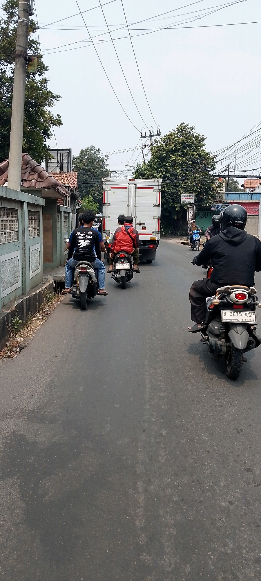 A wide-angle, vertical, low-angle shot captures a busy street scene in Bekasi, Indonesia. The asphalt road is lined with a grey, textured concrete wall on the left, partially obscured by lush green trees and a tiled roof. Power lines crisscross the sky, casting shadows. Several motorbikes are in motion, with riders wearing casual clothing and helmets. In the foreground, a person on a motorbike wearing a black hoodie and helmet is riding away from the camera. Ahead, another motorbike carries two passengers, one in a red jacket. Further down the road, a white box truck is visible. The general atmosphere is one of daily life on a city street.