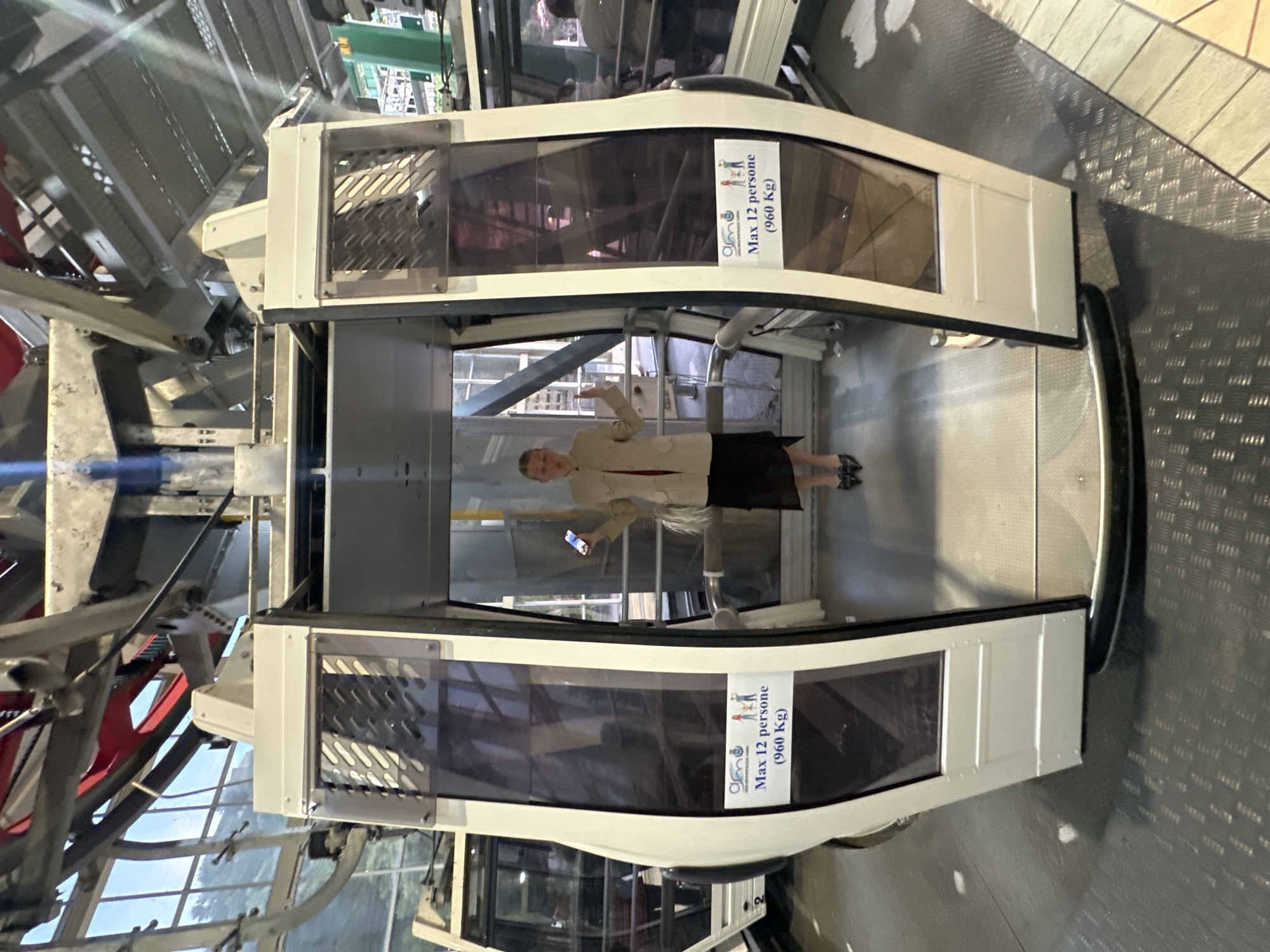 The image captures the interior of a cable car or gondola cabin, seemingly at a station, rotated ninety degrees clockwise. One person, a woman, is the primary subject, standing centrally within the cabin. She wears a light-colored jacket over a dark top and skirt, with dark shoes, holding a smartphone in her left hand and her right arm slightly raised, possibly posing for a photo or adjusting something.
The cabin itself is modern, featuring large, tinted windows framed in white or light-colored material, and a metallic floor. Surrounding the cabin, the intricate machinery of the cable car system is visible, including metal girders, tracks, and a large red wheel partially seen on the left. The floor outside the cabin is a textured metal platform, transitioning to tiled flooring further into the station.
On the cabin windows, two identical white signs are affixed, both stating "Max 12 persone (960 Kg)", accompanied by a small graphic of three stylized people. This indicates the capacity limit of the cabin. The lighting suggests it is daytime, possibly late afternoon or early evening, with some light filtering in from outside through the mechanical structures. The scene conveys a moment of boarding or waiting within the cable car system, likely in Taormina, Italy, given the provided context.