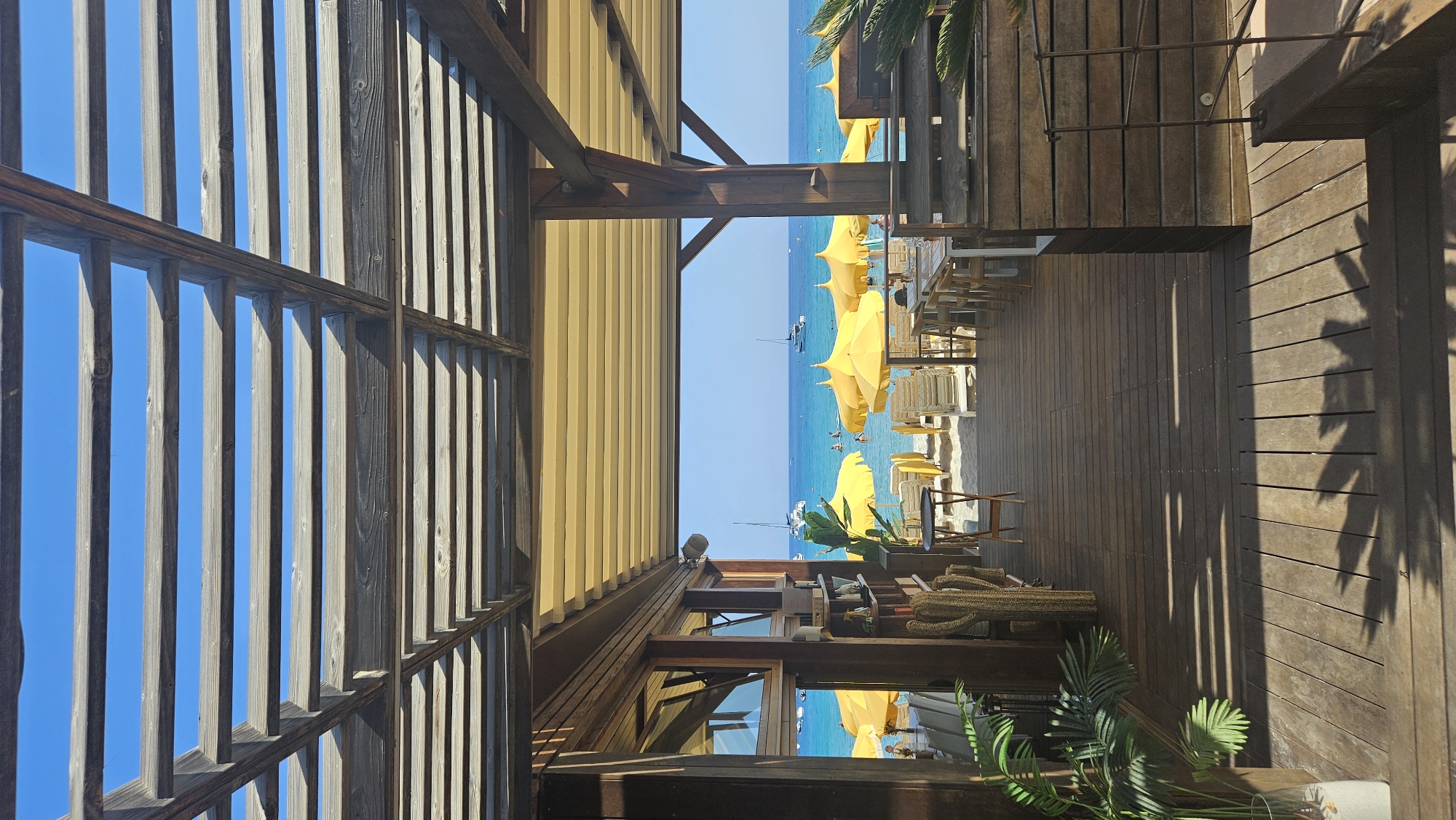 This is a photograph taken from the covered wooden deck of a beachside establishment, likely a restaurant or bar, in L'Île-Rousse, France. The viewpoint looks out towards the Mediterranean Sea.
In the foreground, a wooden deck extends towards the beach, with shadows of palm fronds cast across it. To the left, a potted green plant in a glass vase is visible. A wooden structure with slatted walls and a metal railing is to the right.
The middle ground reveals the beach area, furnished with rows of white lounge chairs and vibrant yellow umbrellas. A wooden walkway leads from the deck onto the sand. Several people are visible on the beach and in the shallow water.
The background is dominated by the clear blue sea and a bright blue sky. Sailboats are dotted across the water. The architecture of the establishment features exposed wooden beams supporting a yellow retractable awning. The overall impression is one of a sunny, relaxed atmosphere at a coastal resort.