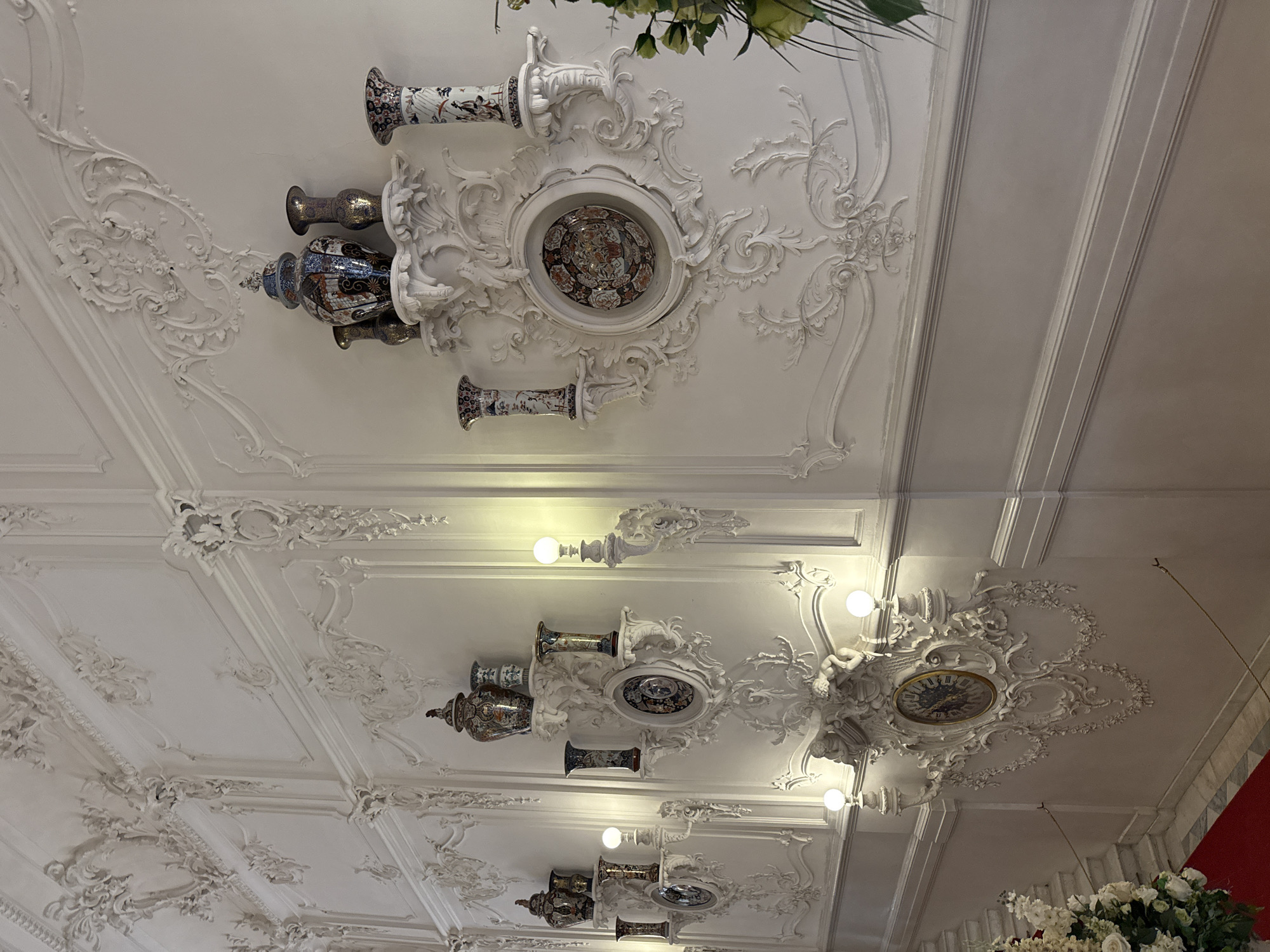 The image depicts a richly ornamented wall in the Catherine Palace, located in Pushkin, Russia. The wall is covered in intricate white stucco work, characteristic of Rococo style, featuring elaborate carvings of floral motifs, scrolls, and cherubic figures. Mounted on decorative shelves are several antique porcelain vases and urns, some with blue and gold decorative patterns. A large, ornate clock is centrally positioned on the wall, surrounded by further stucco embellishments and flanked by wall sconces with glowing light bulbs. To the left, a portion of a staircase with a red carpet runner is visible, along with a large floral arrangement. The scene is brightly lit, highlighting the detailed craftsmanship of the palace's interior. There are no people visible in the image.