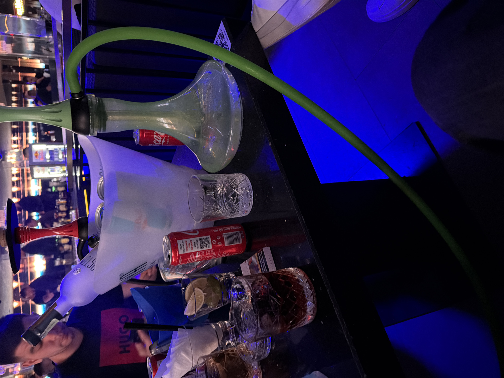 The image captures a dimly lit, social setting, likely a bar, club, or lounge, seen from a slightly elevated perspective. The photograph is oriented vertically, showing a table surface laden with various items in the foreground and midground, while the background features prominent blue artificial lighting.
In the foreground, a light green hookah with a long, flexible green hose dominates the left side. Beside it, an ice bucket contains several items, including at least two Red Bull cans and a bottle of what appears to be vodka, featuring a distinctive shape and possibly the word "PREMIUM VODKA" visible on its label. Multiple glasses, some with an ornate crystal-cut design, are scattered across the table. Some glasses contain a dark amber liquid with ice and lime wedges, while others are empty. Several red Coca-Cola cans are also visible; one prominently displays a QR code and Cyrillic text, including "ОРИГИНАЛЕН ВКУС" (Original Taste), indicating a Bulgarian context. A pack of cigarettes lies next to a glass, and crumpled white napkins are also present.
A person, possibly a male, is partially visible in the lower left portion of the image, wearing a dark t-shirt with the red "HUGO" logo printed on it. The background on the right is bathed in a vibrant blue light, suggesting LED or neon illumination, creating a modern and atmospheric ambiance. Other blurred lights and reflections in the deeper background hint at the presence of more patrons or elements of the venue. The overall scene depicts an evening or nighttime gathering focused on socializing, drinking, and smoking hookah.