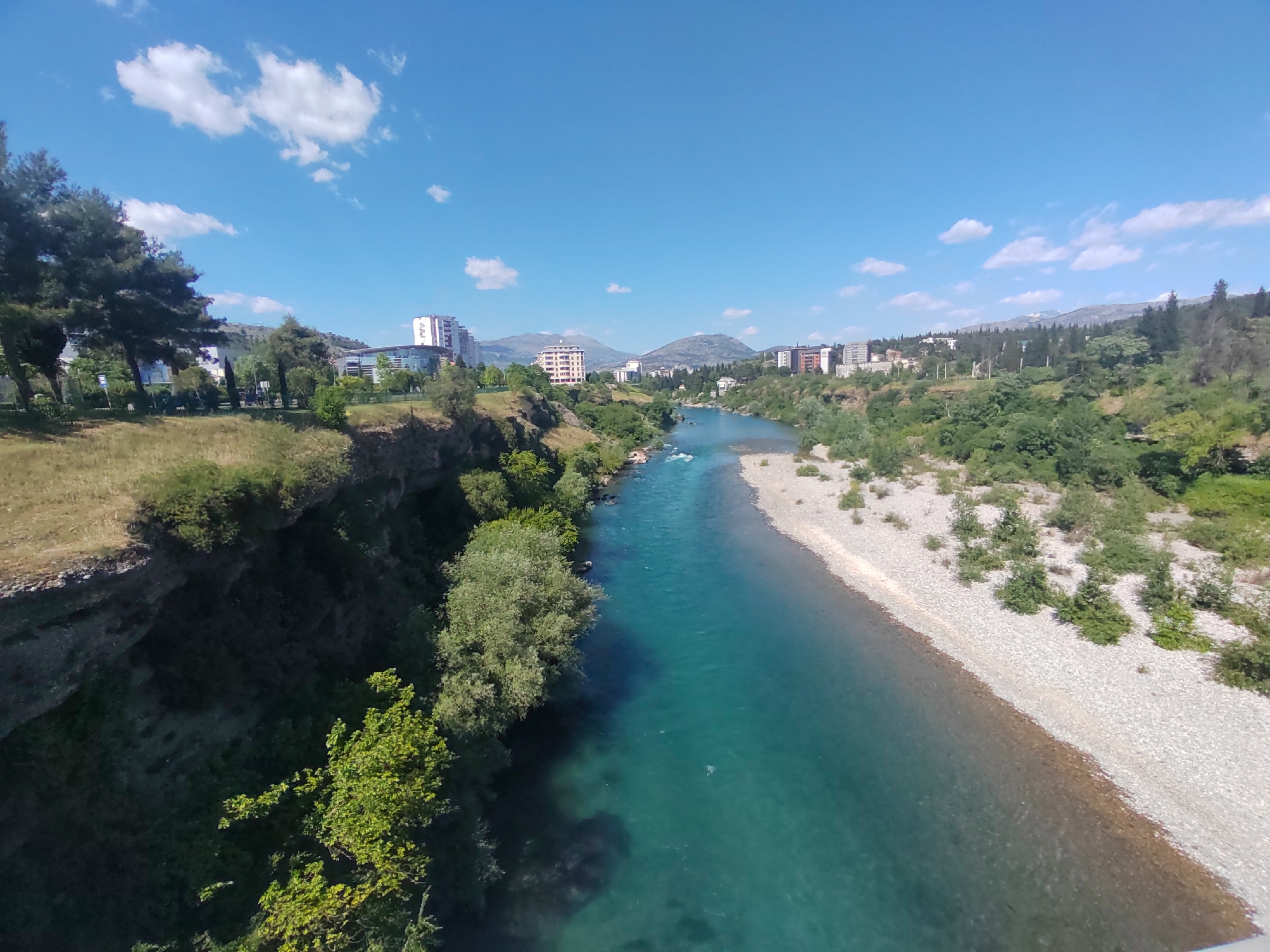 The image depicts a river flowing through the city of Podgorica, Montenegro, likely taken from a bridge. The river is a vibrant turquoise color, and its banks are lined with greenery on one side and a gravelly, stone-covered bank on the other. In the background, buildings and mountains can be seen, suggesting an urban environment. The sky is blue with scattered clouds, indicating a sunny day.