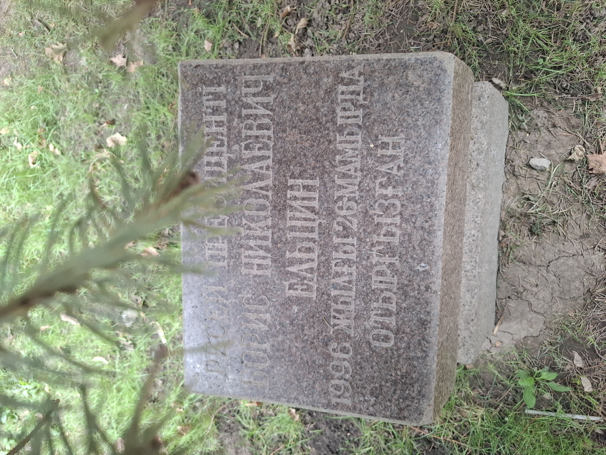A granite monument with engraved text is set in a grassy area under a coniferous tree in Almaty, Kazakhstan. The monument is on a concrete base, and the surrounding ground is covered in grass and dirt. The engraved text appears to be in Cyrillic and may be a memorial.