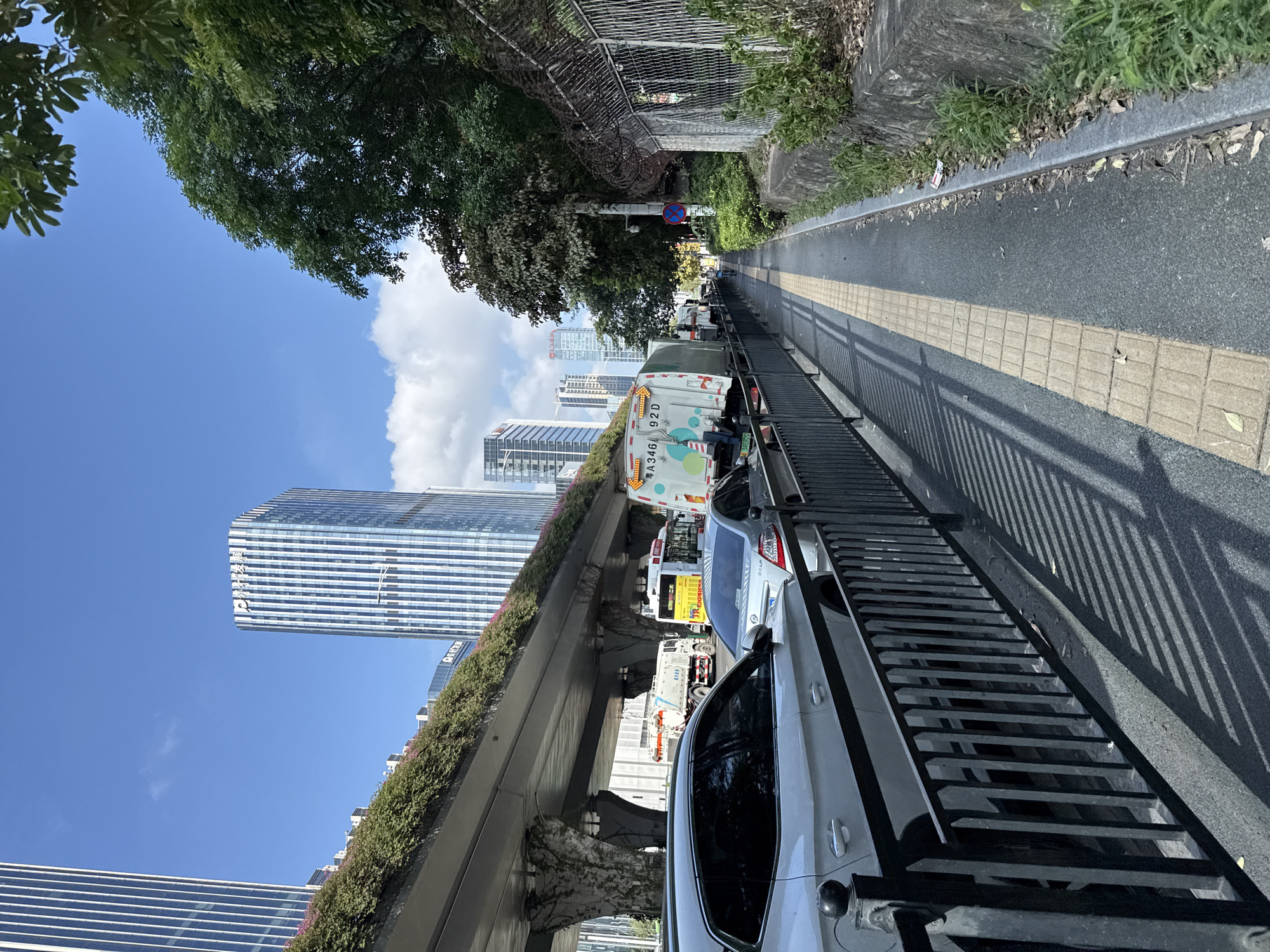 The image depicts a city street scene in Huangpu District, China. The foreground features a black metal railing along a sidewalk. Behind the railing, several cars are parked or in traffic, including a silver sedan and a white SUV. A sanitation truck with colorful graphics is visible further down the street, as is a bus. In the background, a multi-level overpass with lush greenery is prominent. Beyond the overpass, modern high-rise buildings with reflective glass facades dominate the skyline, under a clear blue sky with scattered clouds. Trees and a chain-link fence with barbed wire are present along the right side of the image, adjacent to the sidewalk. The environment suggests an urban setting with ongoing traffic and infrastructure.