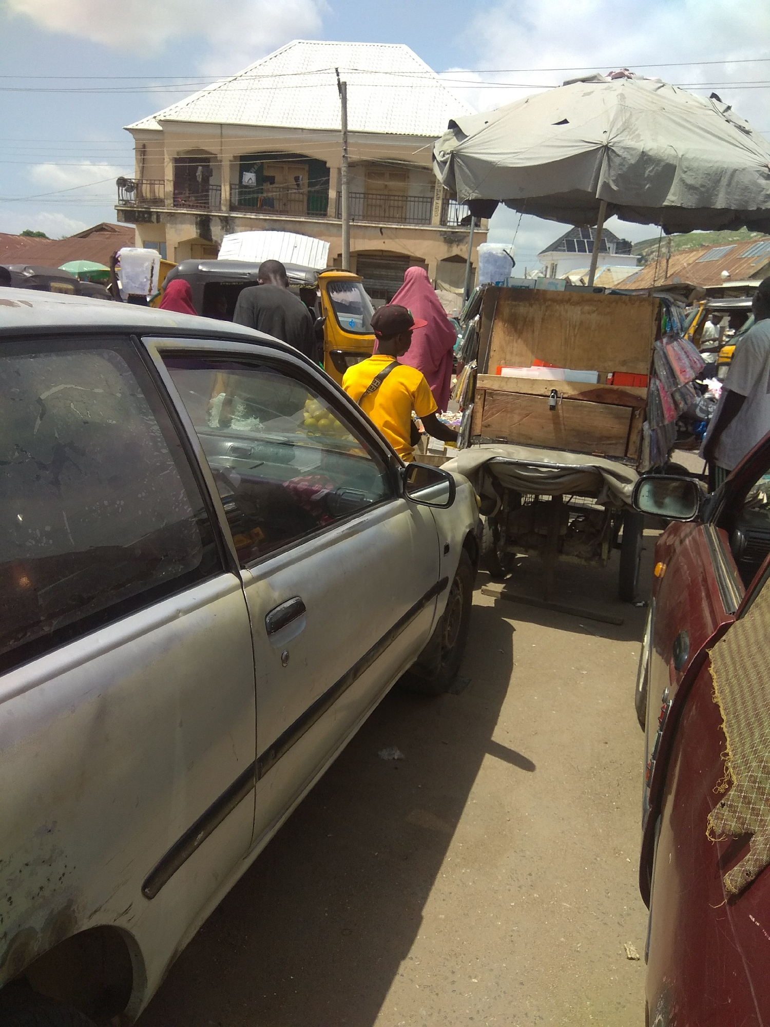 The image depicts a street scene in Jalingo, Nigeria. In the foreground, a light-colored sedan is parked, facing left. To its right, a red vehicle is partially visible, also parked. Between the vehicles, a wooden cart is positioned. The cart is covered with a tarp and appears to be laden with goods, including what look like stacked crates or boxes. Behind the cart, a yellow auto-rickshaw is also present.
Several individuals are visible in the scene. A person wearing a yellow shirt and a dark cap is standing near the cart, facing away from the camera. Another person in a dark shirt is visible in the yellow auto-rickshaw, also facing away. Further back, a person in a pink garment is standing to the left of the cart, and another person in a white shirt is standing behind the cart.
In the background, a two-story building with a white tiled roof and balconies is prominent. The building appears to be a commercial establishment or residential complex. Other buildings with corrugated metal roofs are visible in the distance, characteristic of urban architecture in Nigeria. The sky is partly cloudy. The ground is paved, suggesting a street or market area. The overall impression is a busy street, possibly a market or transport hub, in an urban setting.