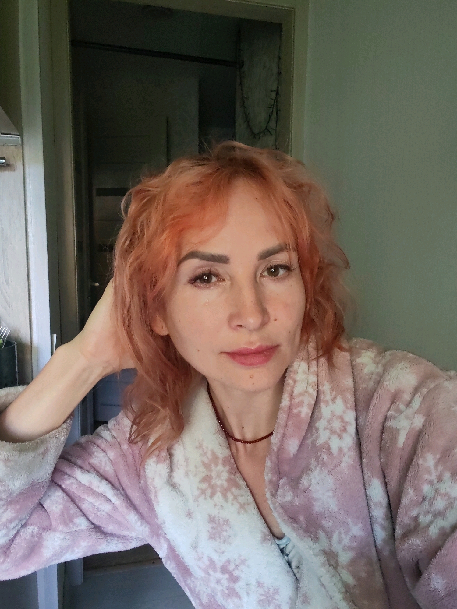 The image is a close-up selfie of a woman with short, wavy, ginger-colored hair, wearing a pink and white snowflake-patterned bathrobe. She has light skin, light eyebrows, and is wearing subtle makeup, including pink lipstick. A thin, dark red beaded necklace is visible around her neck. Her left hand is resting on the back of her head, and a teardrop is visible on her right cheek. The background is dimly lit, suggesting an indoor setting, possibly a home. The environment appears to be an apartment in Chaikovsky, Russia, with a doorway and a wall with some decorative string lights visible.