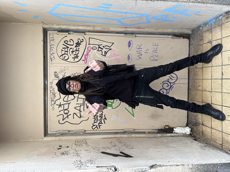 A person with dreadlocks, a black beanie, glasses, and dark clothing stands in front of a door covered in graffiti, including the words "WAR IS PEACE." They are holding up both middle fingers to the camera. The scene appears to be an urban alleyway or passage with graffiti on the walls. The overall tone is rebellious.