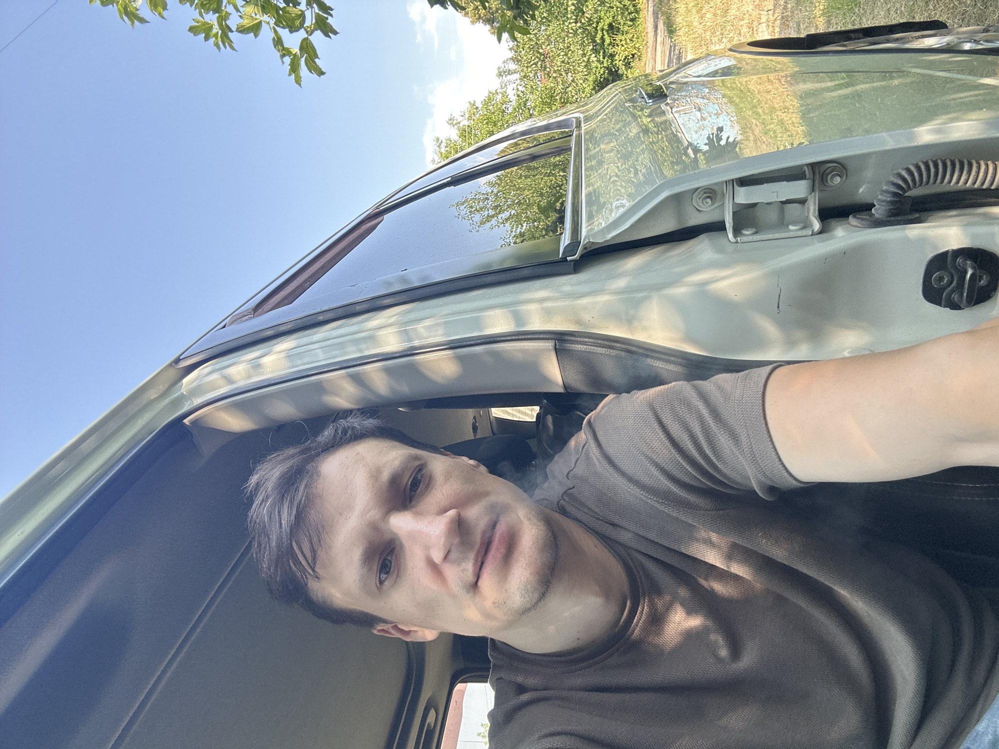 A man with short, dark hair and a light complexion is seated inside a vehicle, looking directly at the camera. He is wearing a dark olive-colored t-shirt. The driver's side door of the vehicle is open, revealing the interior and the man. The car appears to be light in color. Sunlight filters through the leaves of trees, casting dappled shadows on the man and the car's interior. The background shows a rural setting with green trees and dry grass under a clear blue sky. The man is positioned in the foreground, with the open car door and the outdoor environment forming the background. This image was taken in Shatava, Ukraine.