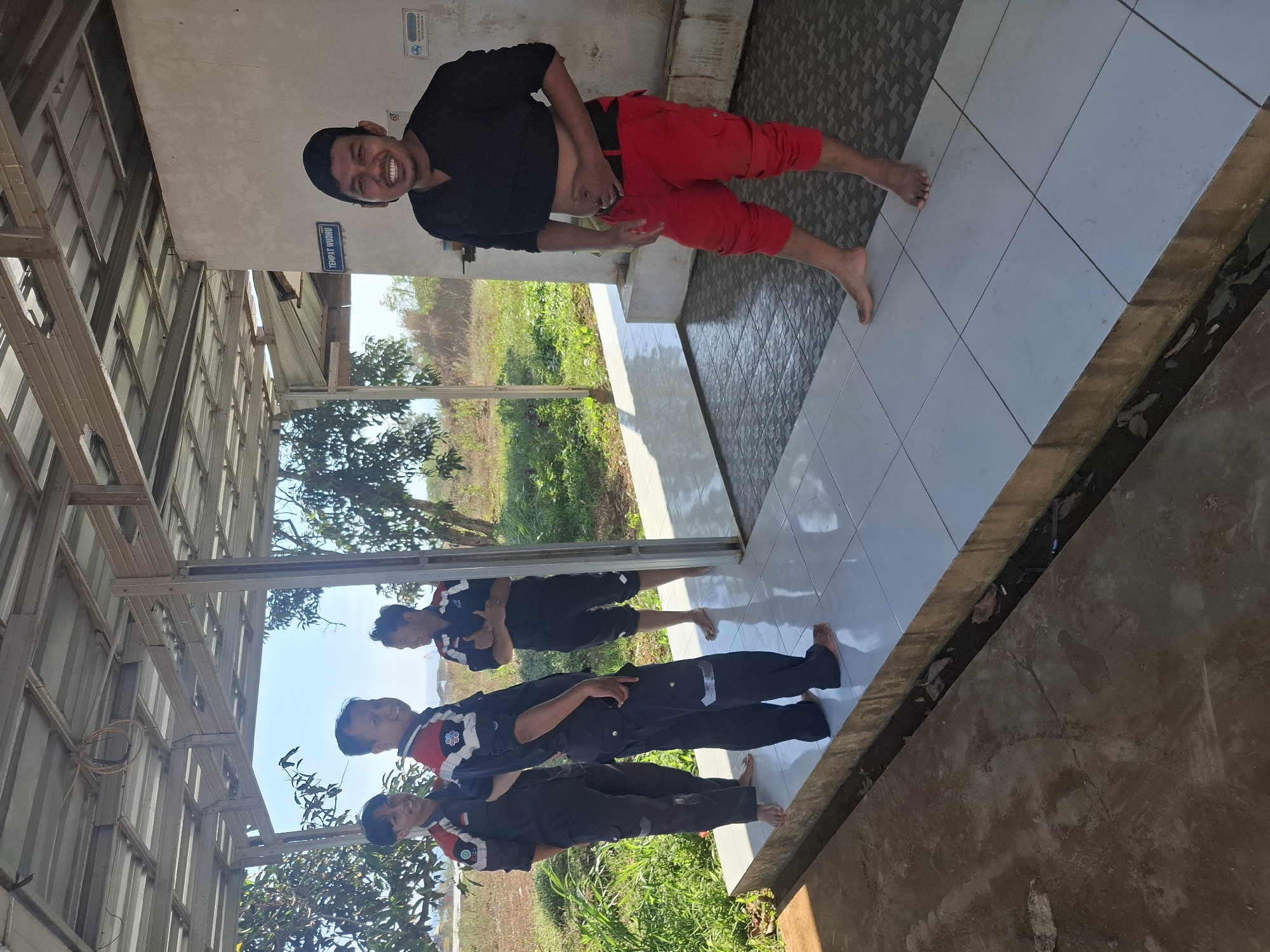 Four men are present in a covered outdoor area with a tiled floor and a corrugated metal roof supported by metal beams. The man on the far right wears a black t-shirt, red pants, and a black cap, and is pulling up his shirt to expose his stomach while smiling and touching his abdomen. The other three men are on the left, wearing matching dark blue uniforms with red and white accents on the collars and sleeves. They appear to be standing and talking. The environment is sunny, with trees and dry vegetation visible in the background. A blue sign on the wall behind the man in red reads "TEMPAT WUDHU," indicating this is a place for ritual washing. The overall setting suggests an outdoor facility, possibly a rest stop or a communal area.