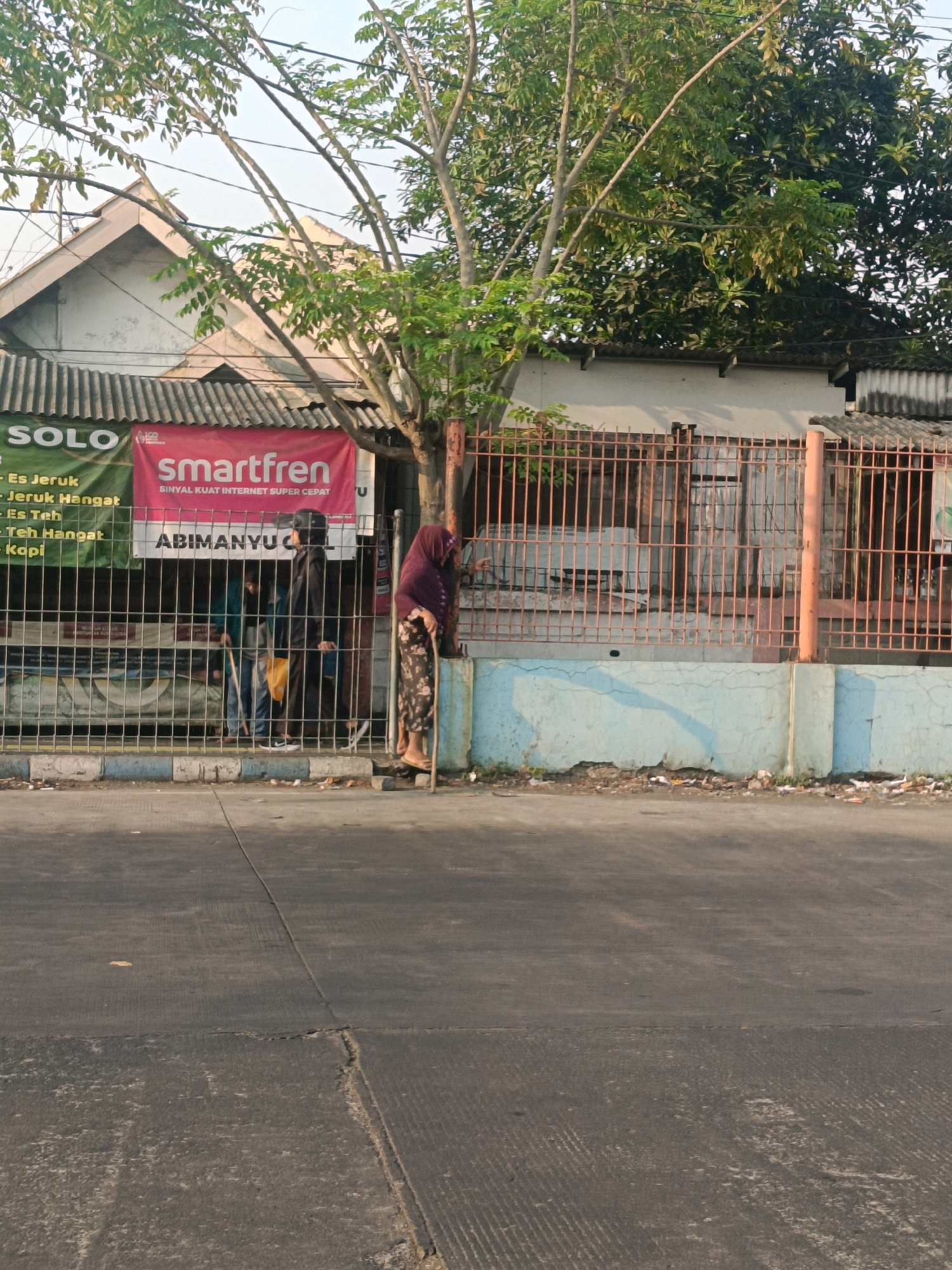 In Kedungrejo, Indonesia, an outdoor scene depicts a street in front of a building with a metal fence. Two individuals, one wearing a dark hijab and patterned trousers, leans on a cane, while another person stands behind the fence wearing a dark hijab and black clothing. A red banner with the Smartfren logo and "SINYAL KUAT INTERNET SUPER CEPAT" is visible on the building, along with other signs indicating products like "Es Jeruk" and "Es Teh". The building has a corrugated metal roof and a light blue concrete wall in the foreground. A tree with green leaves grows near the building. The street in the foreground is paved with asphalt.