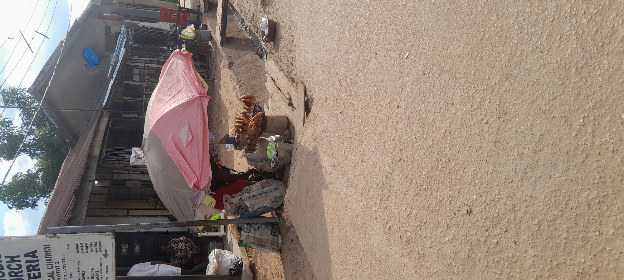 The image shows a street scene in Abuja, Nigeria, with a vendor selling grilled corn under a pink and beige umbrella. The vendor, wearing a red shirt, is partially obscured by the umbrella and is seated on the ground. In front of the vendor are several large bags, possibly containing grains or other goods, and a metal bucket. A metal rack holding several ears of grilled corn is positioned on top of a cylindrical object, which could be a stool or a container, and appears to be a cooking station. The ground is sandy and uneven, with some debris scattered around.
To the left of the vendor, a sign for a church is visible, with some of its text legible, indicating "CHURCH NIGERIA", "CAL CHURCH", and "WEEKLY ACTIVITIES" with times listed for services. A person wearing white is standing near the sign, and another person in a patterned top is seated. A large white bag and a black bag are placed near the entrance of a building behind the seated person.
In the background, buildings with corrugated iron roofs are visible. One building has metal bars on its windows. A blue satellite dish is mounted on the roof of one of the structures. Further in the background, trees are present, and power lines stretch across the scene. The overall environment suggests a market or a roadside commercial area.