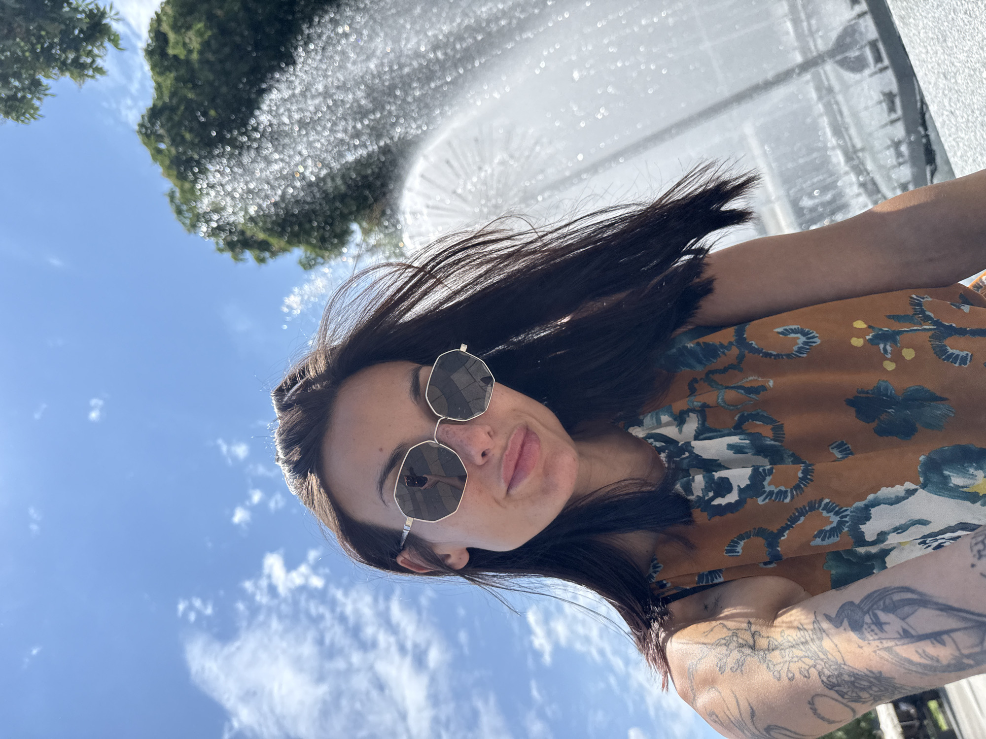 A woman takes a selfie in front of the Pleven Water Cascade fountain in Pleven, Bulgaria. The woman has dark hair and wears sunglasses and a floral top. Tattoos are visible on her arm. The fountain sprays water upwards behind her, and the sky is blue with scattered clouds. A tree is also visible in the background.