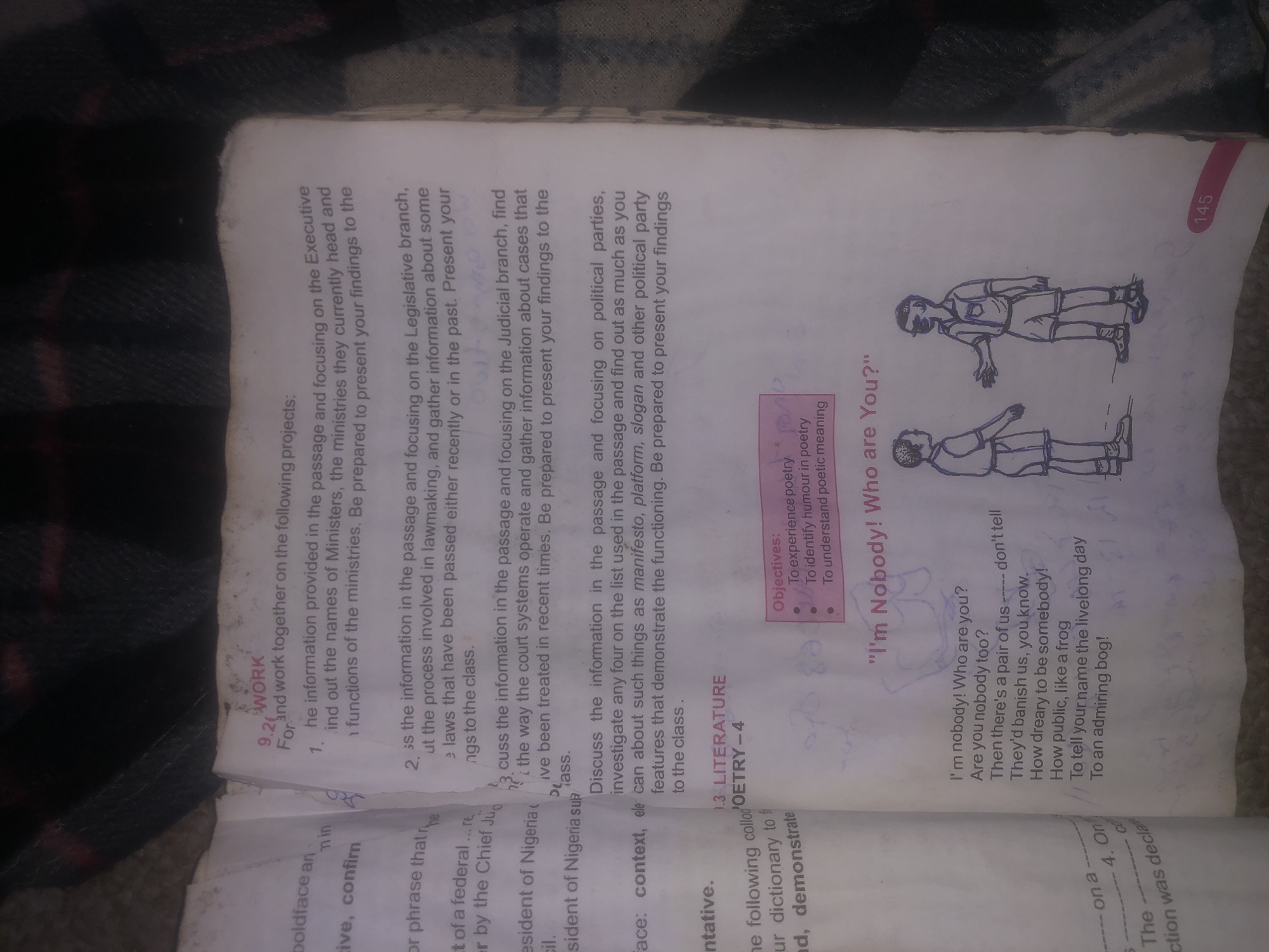 The image is a top-down, close-up view of an open book. The book's pages are filled with text and illustrations, indicating it is likely a textbook or study material. The text is in English and appears to be a mixture of educational content, likely related to civics or social studies given the headings "WORK" and "LITERATURE", and possibly a poem titled "I'm Nobody! Who are You?". One page features a line drawing of two young boys in a conversational stance. The book is lying on a dark, textured surface that could be a blanket or fabric. There are no discernible features within the image that would allow for identification of its location in Jalingo, Nigeria.