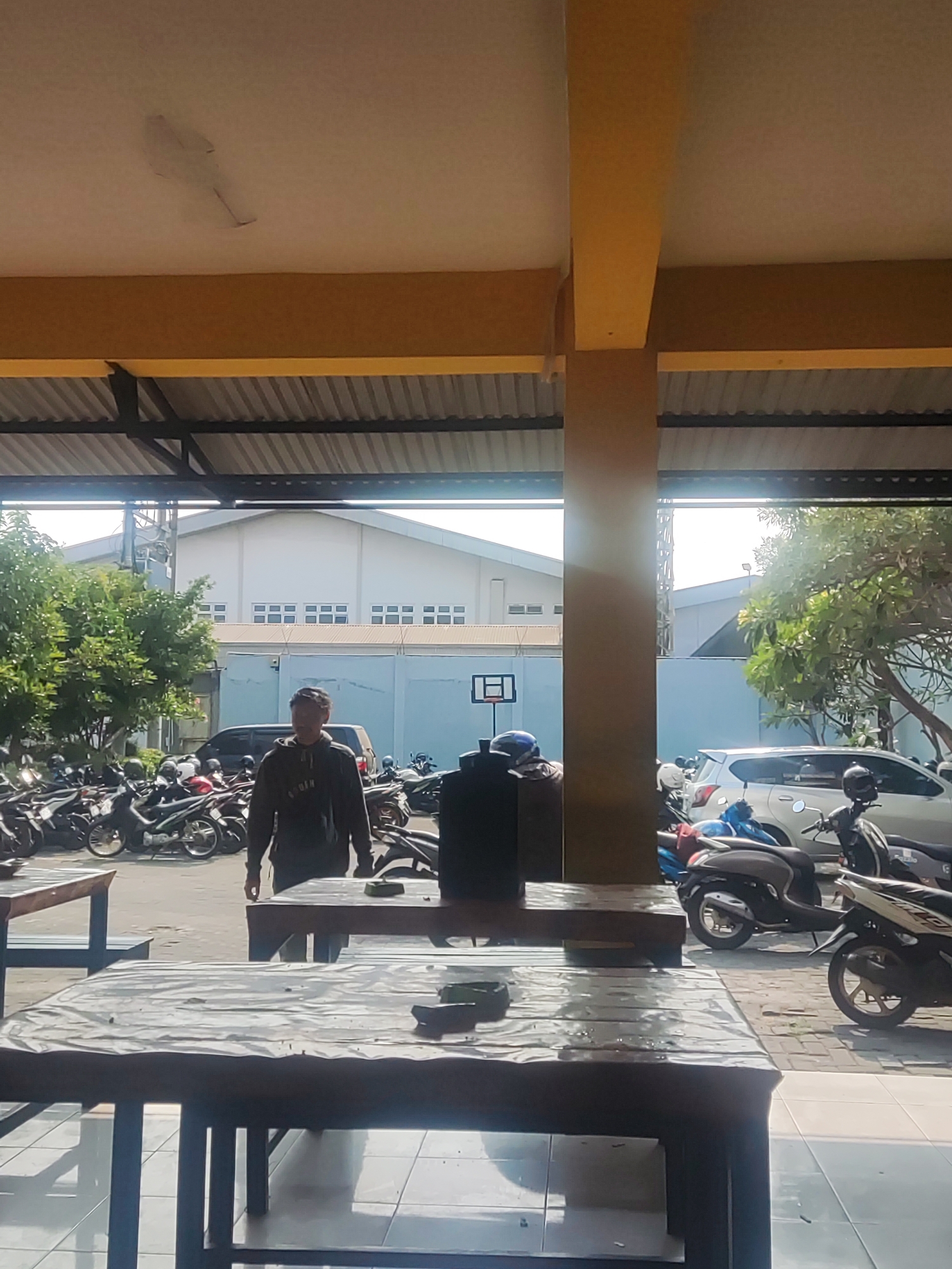 The image shows an outdoor seating area with two tables in the foreground. The tables are dark wood with metal legs. The table on the right has a dark object on it. In the middle ground, a man in a grey hooded sweatshirt is standing. Behind him, there is a parking lot filled with motorcycles and a silver car. In the background, a large white building with rows of windows is visible, along with a basketball hoop and net. Trees and a light pole are also present in the scene. The overall environment appears to be an urban or semi-urban setting in Trihanggo, Indonesia. The lighting suggests it is daytime.