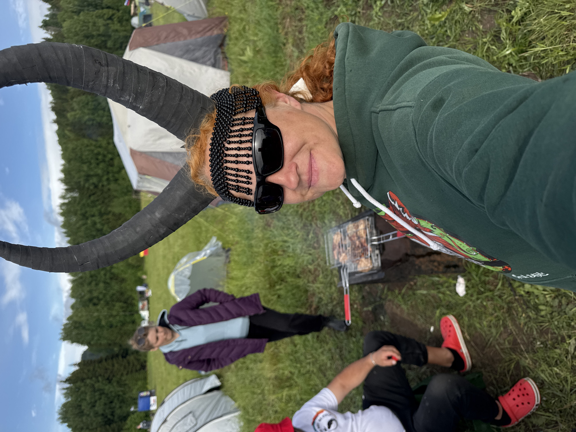 This image captures an outdoor gathering, likely a camping or festival scene, oriented as if rotated 90 degrees clockwise. The setting is a grassy field with a line of trees in the background, under a blue sky with some scattered clouds.
The main subject, a person with auburn hair, is taking a selfie. They are wearing a dark green hooded sweatshirt with a colorful graphic (partially visible) and black sunglasses. A distinctive black beaded headpiece, resembling a net or chainmail, covers their forehead. Attached to their head are large, dark, curved horns, which appear to be wrapped in black tape. The person has a faint smile, suggesting a relaxed mood.
In the mid-ground, a small portable barbecue grill is set up on the ground, with food (possibly meat) cooking and emitting light smoke.
Two other individuals are visible. One person, with light brown hair, stands further back, wearing a purple jacket over a light blue top and dark pants. Another person is sitting on the grass in the foreground, wearing a white t-shirt with a logo, black pants, and bright red Crocs shoes.
Several camping tents of different colors (grey, brown, light green) are pitched across the grassy area in the background, indicating a campsite. The overall scene depicts a casual daytime outdoor social event. Visible text "Real Style" can be seen on the main subject's hoodie.