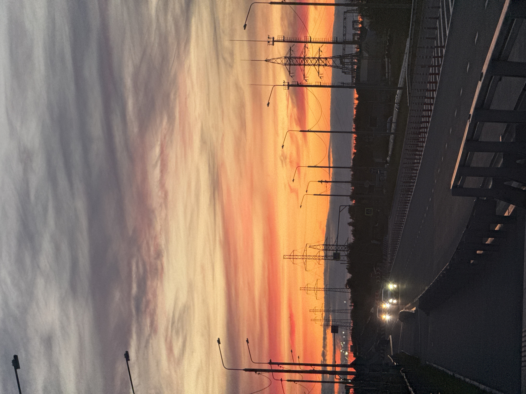 The image captures a vibrant sunset over an urban landscape, likely on the outskirts of Salekhard, Russia. A highway is visible in the foreground, with two cars driving with their headlights on, indicating approaching dusk. Streetlights line the road, and a pedestrian walkway is to the left. In the background, several tall electrical towers stand silhouetted against the colorful sky, which is ablaze with hues of orange, pink, and purple. The horizon is marked by a dark line of trees and possibly some industrial structures. The overall atmosphere is one of transition from day to night, with the dramatic sky providing a striking backdrop to the infrastructure.