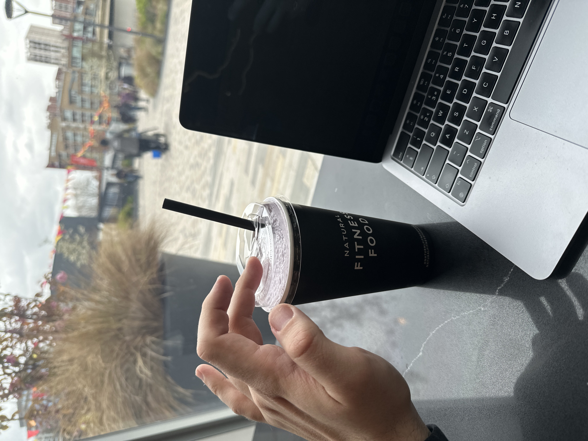 Here is a description of the image:
The image shows a person's hand holding a black smoothie cup with a straw. The cup is from a brand called "Natural Fitness Food." Next to the cup is a partially visible silver laptop, showing part of the screen and keyboard. In the background, visible through a window, is an urban scene with buildings, trees, and people walking. The scene suggests an outdoor area, possibly a waterfront promenade. The angle of the photo implies it was taken at a table, probably indoors.