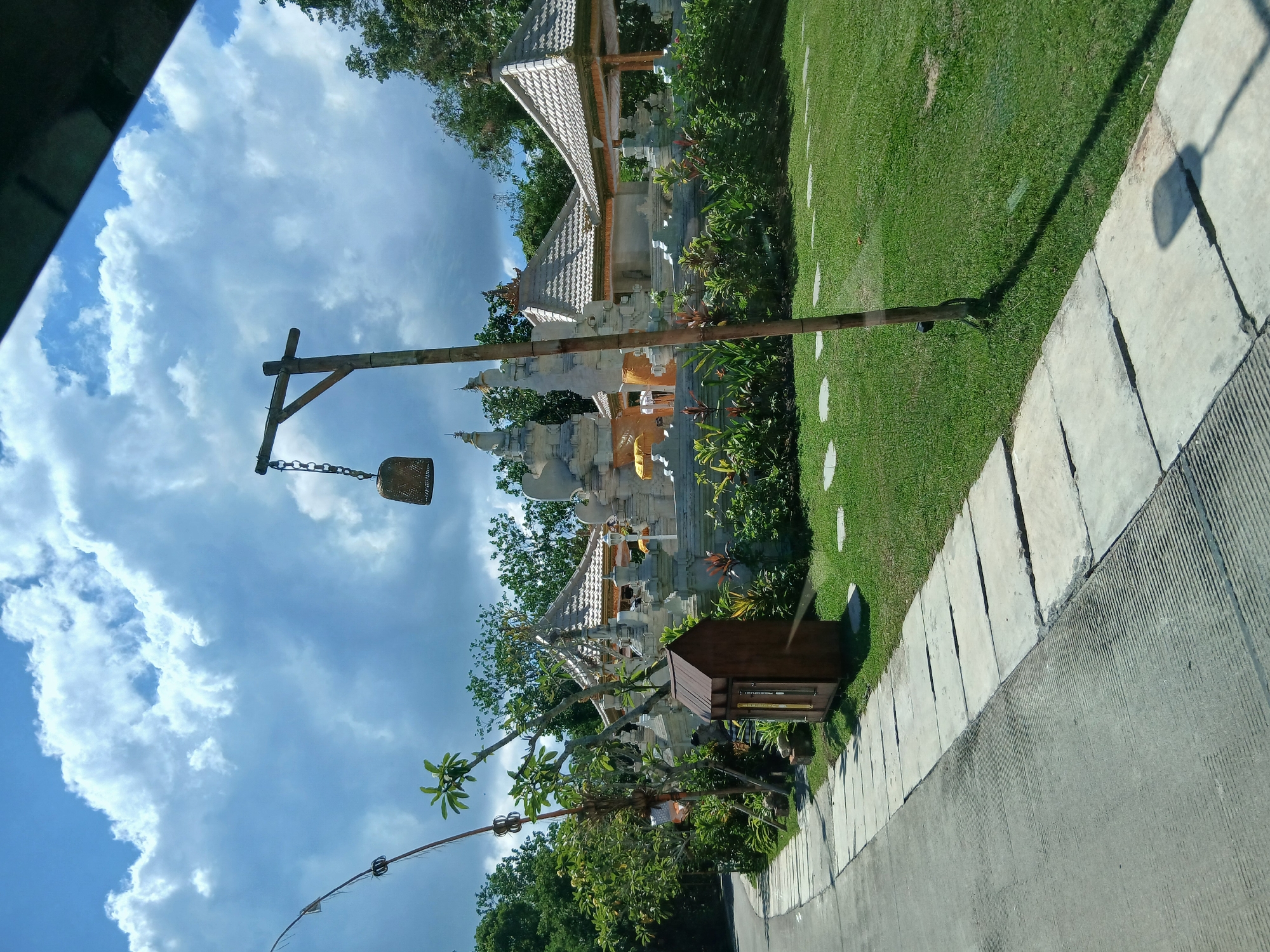 The image depicts a pathway leading towards a Balinese-style temple complex in the city of Mengwi, Indonesia. The pathway is paved with concrete slabs and bordered by a manicured green lawn. To the left of the pathway, there is lush vegetation, including trees and ornamental plants. A tall wooden pole with a hanging bell is positioned near the center of the image, casting a shadow on the lawn. The temple complex itself features ornate white stone structures with tiered roofs, adorned with decorative elements and yellow fabric. The background is filled with dense green trees and a bright blue sky with scattered white clouds. No people are visible in the image.