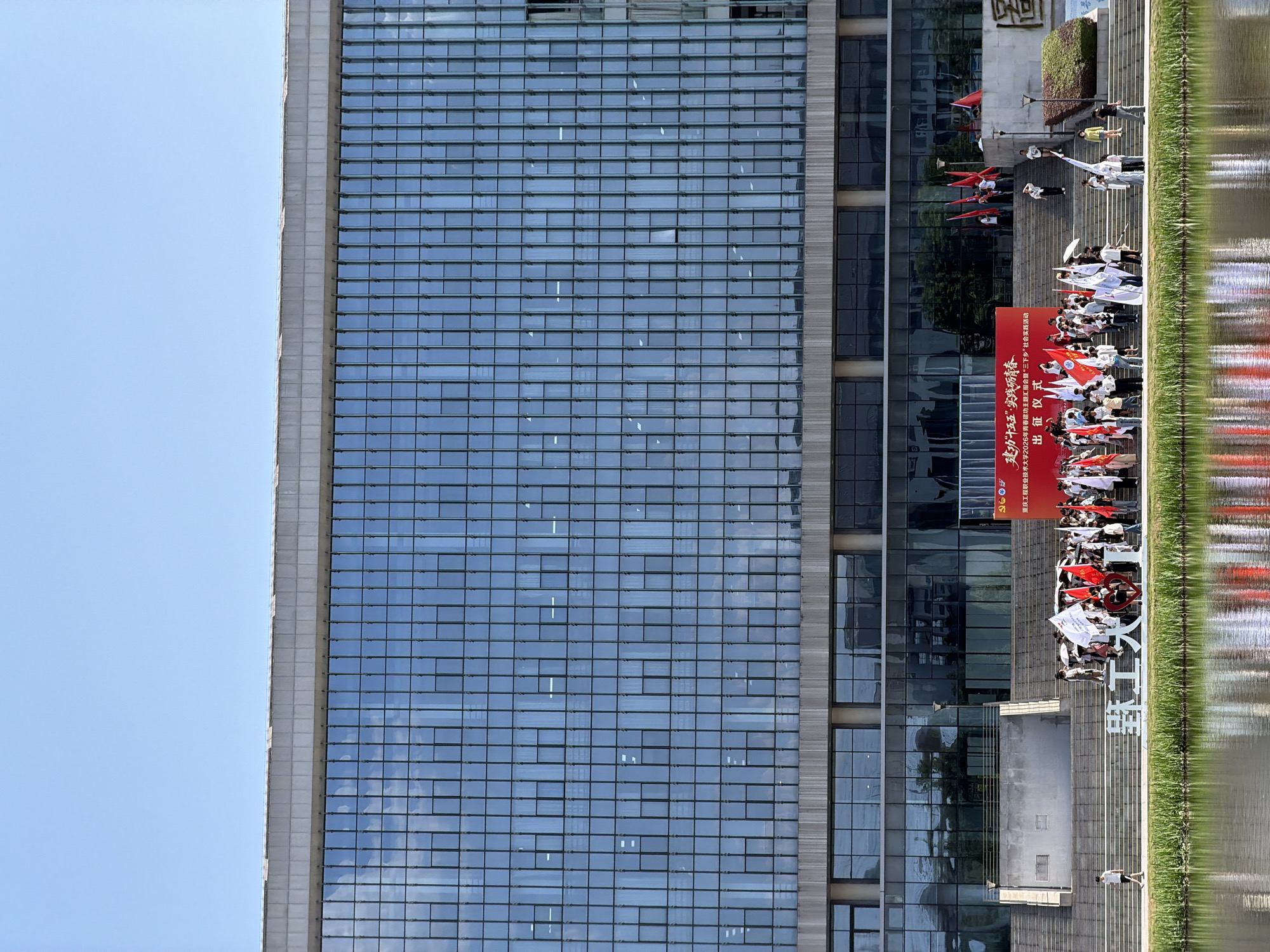 This is a medium-shot, eye-level outdoor photograph taken on a sunny day in front of a modern glass-facade building. The building, likely an educational institution, is a dominant feature, with a reflective glass exterior. In the foreground, a calm body of water with visible reflections of the sky and surrounding greenery stretches across the bottom of the frame. A strip of lush green grass and reeds separates the water from a concrete embankment and a wide set of stairs.
On the stairs and the embankment, a group of people, likely students, are gathered. Many are holding white and red flags, suggesting a formal event or celebration. Some individuals are walking up and down the stairs, while others are standing in groups. A large red banner with Chinese characters is visible on the right side of the stairs, indicating the purpose of the gathering.
To the left of the stairs, a large white sculpture with Chinese characters, possibly the name of the institution or a motto, is displayed prominently. A lone individual is walking on the embankment to the left of the sculpture. The sky above is clear blue with no clouds. The scene appears to be a campus or public space in the city of Jiangjin, China, specifically at the Jiangjin Vocational College, judging by the prominent placement of the college's name on the sculpture and the banner.