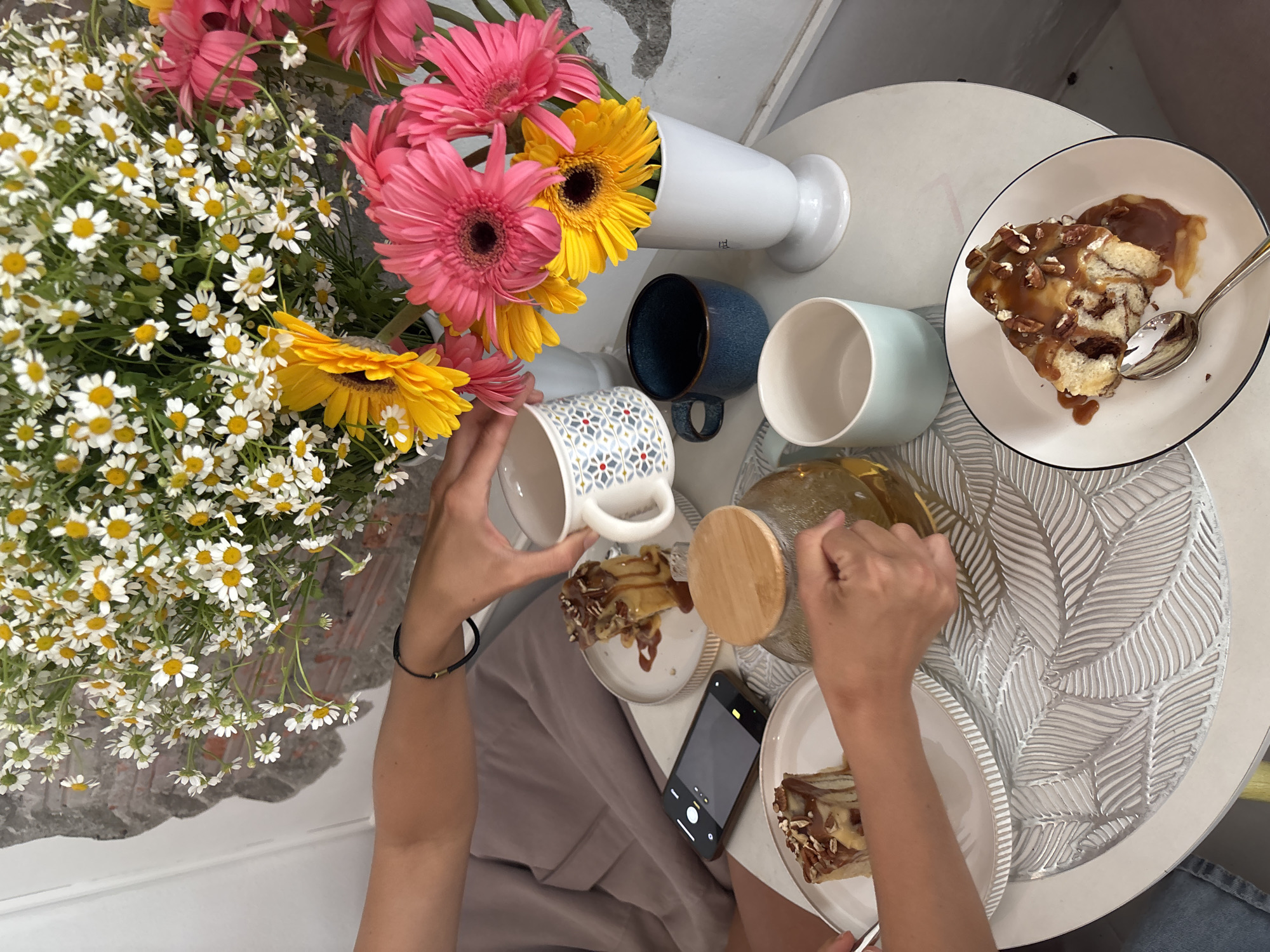 Overhead shot on a white table with a leaf-patterned surface. A bouquet of white daisies, pink gerberas, and yellow sunflowers sits in a white vase. A second vase of the same flowers is to the right. Three cups of various colors sit between the vases and a plate with a dessert with caramel and nuts. A person with a bracelet is holding a patterned mug over a plate of similar dessert, next to a glass pitcher. A phone is on the table between the plates. The scene appears to be set indoors. The city location Phuket City Municipality, Thailand is not explicitly evident from the provided image.