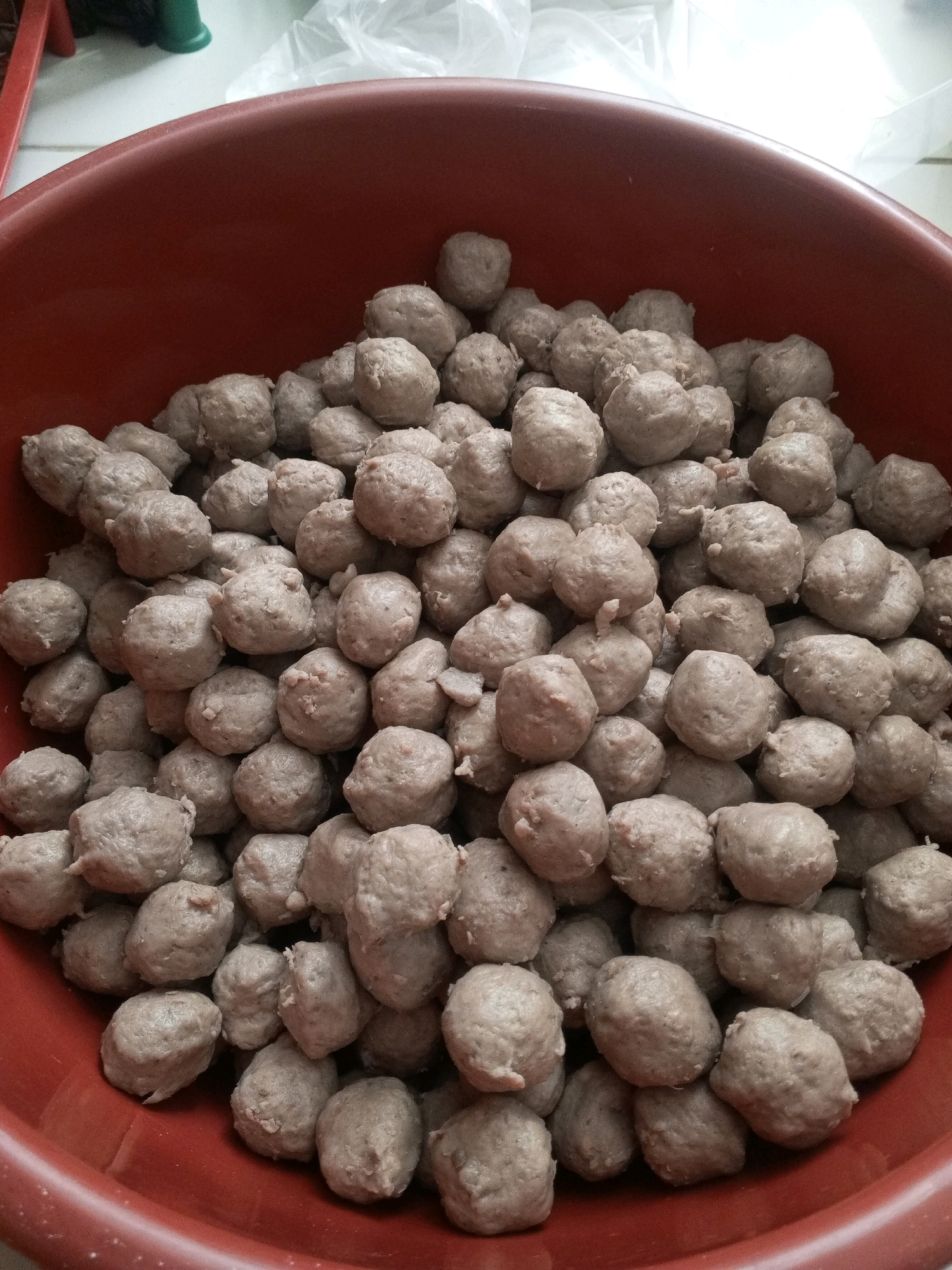 A close-up, overhead view captures a large, round, dark red plastic bowl overflowing with numerous small, spherical meatballs. The meatballs are a pale brown, with a slightly textured, uneven surface, suggesting they are uncooked. They are densely packed, filling the bowl and creating a mound that spills over the rim in some areas. The lighting is overhead and slightly diffused, casting soft shadows and highlighting the individual shapes of the meatballs. The background is out of focus, showing what appears to be a white plastic bag and a hint of a green object. The overall scene suggests a food preparation or storage setting, possibly in a home or market. This image was taken in Rangkasbitung, Indonesia.