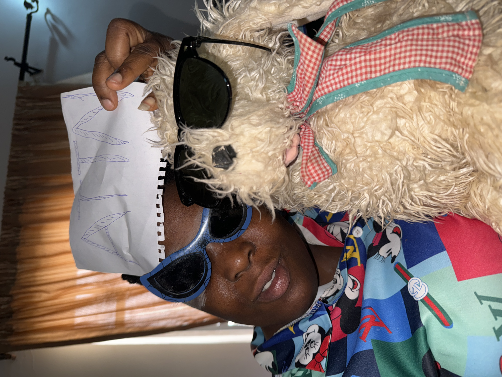 A person is holding a piece of paper with illegible writing next to their face. The person is wearing blue framed sunglasses and a colorful shirt with cartoon characters and logos. A stuffed animal with sunglasses and a checkered bow is resting on the person's shoulder. The background includes a partially visible brown curtain. The photograph was taken indoors. There is no indication of the location being Sabon Tasha, Nigeria.