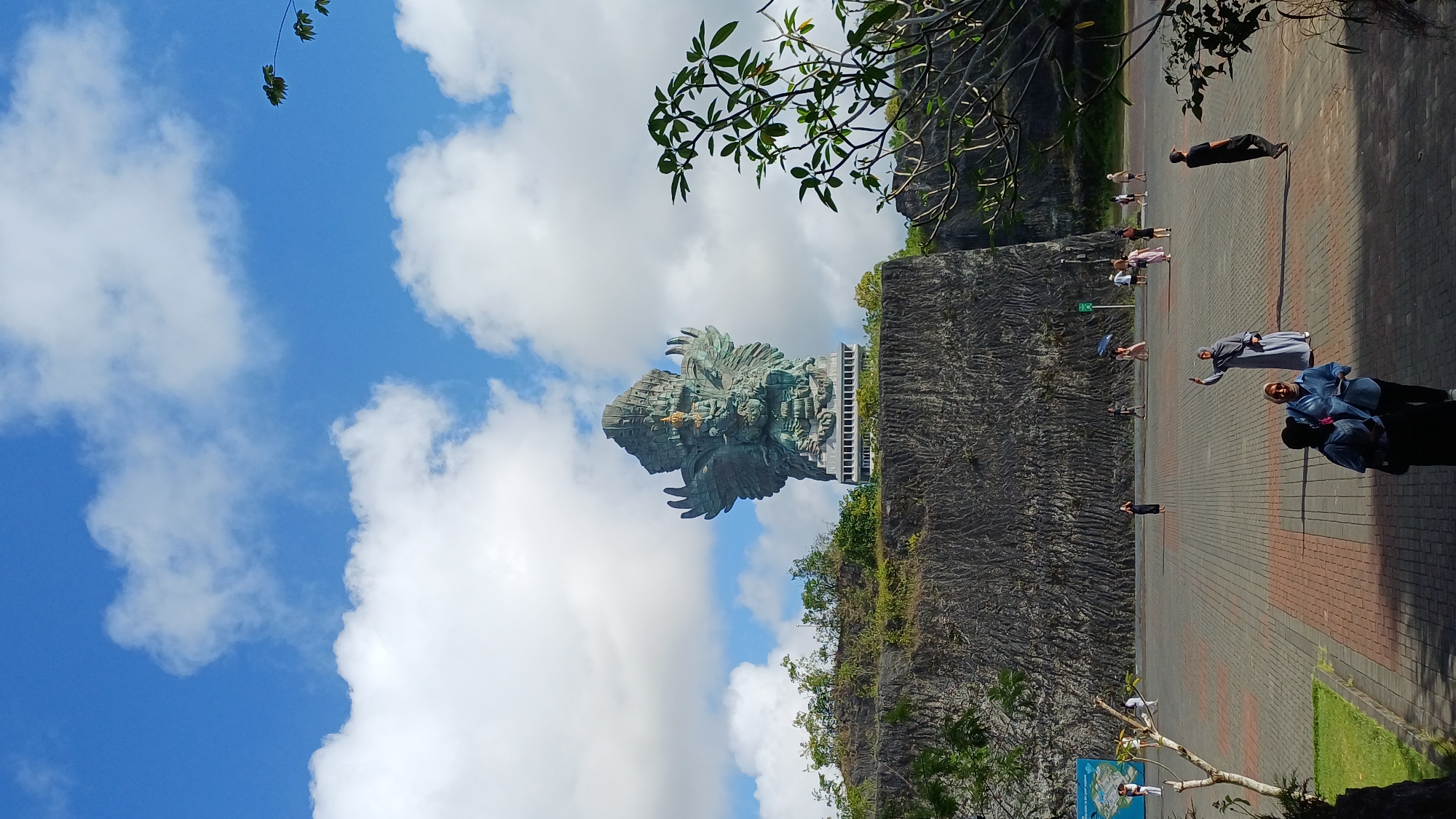 The image shows the Garuda Wisnu Kencana statue in Kuta Selatan, Bali, Indonesia, set against a blue sky with scattered white clouds. The statue is partially framed by green foliage in the upper sections of the image. A group of people are walking on a brick-paved area near the statue’s base, indicating its role as a tourist attraction. The surrounding area features a textured, rocky landscape.