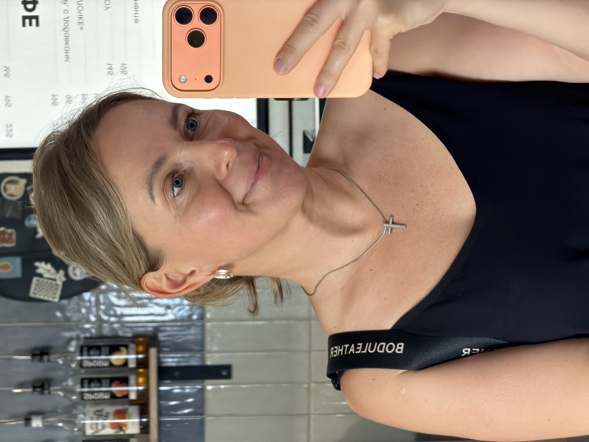 A woman is taking a selfie in what appears to be a cafe. She has blonde hair and is wearing a black tank top and a silver cross necklace. She is holding a peach-colored phone. In the background, there are bottles of syrup on a shelf and a tiled wall. The phone's case is partially visible, and the woman's left shoulder strap has the text "BODETEHTHEB" printed on it. The cafe's sign, written in Cyrillic, is visible in the background, and based on the context provided by the text "city of Chaikovsky, Russia", this scene is likely taking place in a cafe in that city.