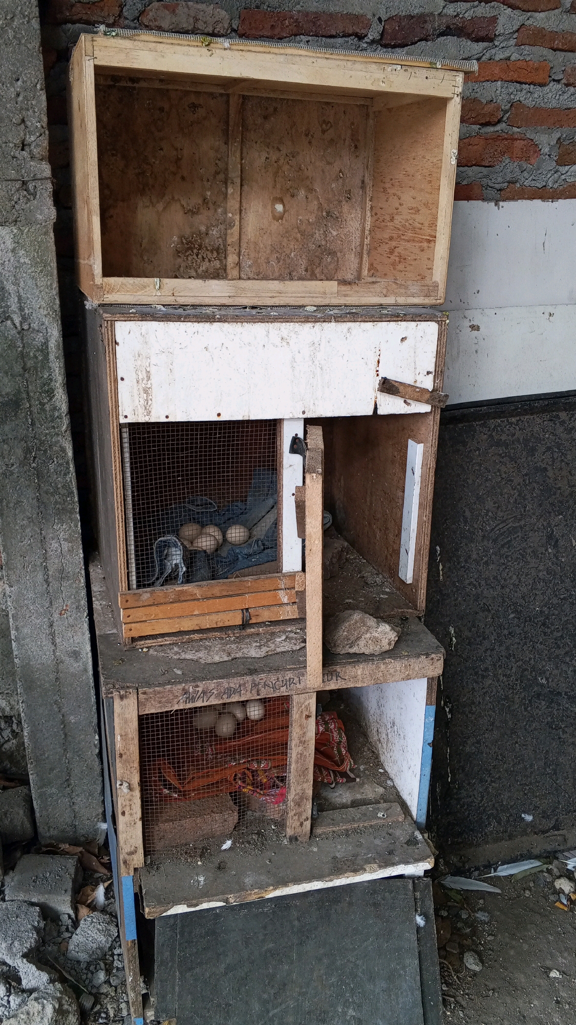 The image shows a stack of three wooden enclosures, possibly for birds or other small animals, against a brick wall in an outdoor setting. The enclosures appear aged and weathered, with some signs of disrepair. The bottom two enclosures have wire mesh fronts, and the middle one contains several eggs and what appears to be a dark blue fabric. The top enclosure is open and appears empty. The ground around the enclosures is littered with debris, including bricks and feathers. The overall impression is one of a utilitarian, possibly abandoned, structure in a somewhat neglected environment. The context of Semarang, Indonesia, suggests a tropical or subtropical climate, which could explain some of the weathering and vegetation.