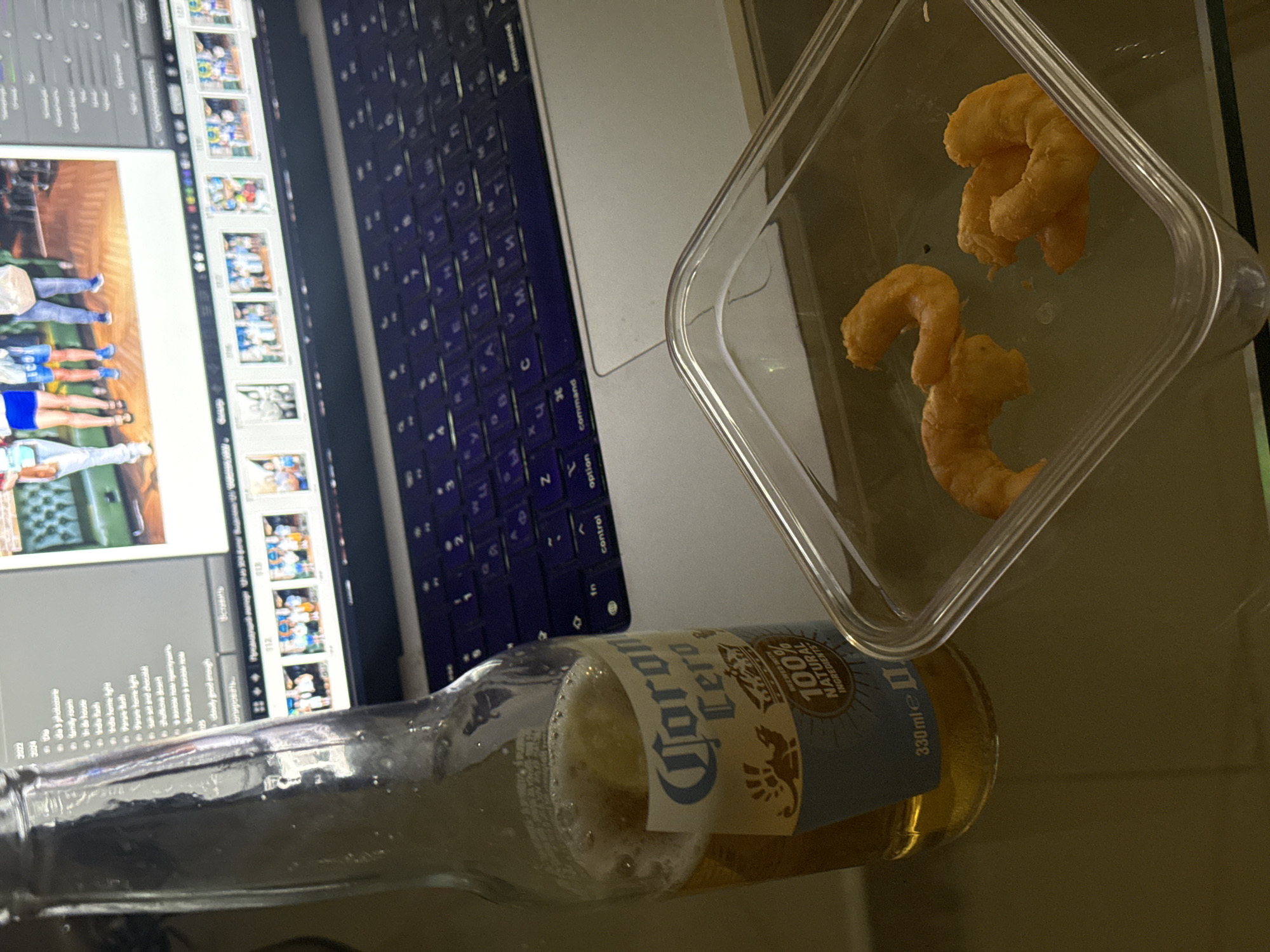On a reflective surface, possibly a glass table, is a partially consumed Corona Cero beer bottle, a clear plastic container with several orange cheese puffs, and a partially visible laptop screen. The laptop display shows what appears to be a photo editing software interface, displaying rows of images that include people in blue uniforms. The laptop keyboard is partially visible.