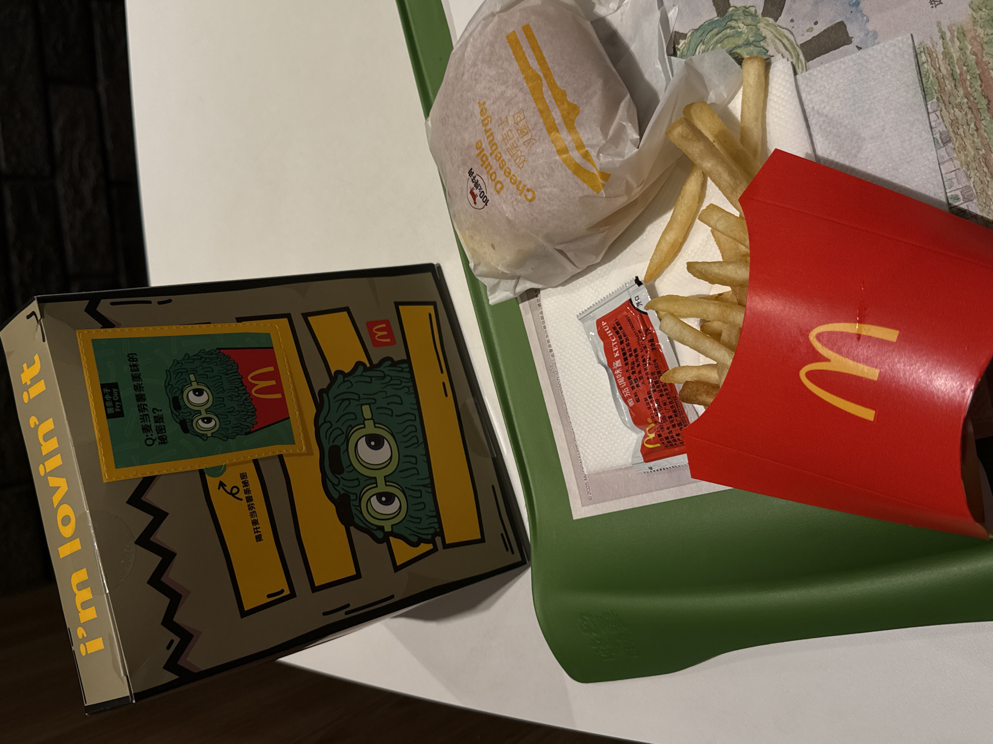 A McDonald's meal is presented on a green tray. In the foreground, a red McDonald's fry carton overflows with golden french fries. To the right of the fries is a wrapped cheeseburger, identifiable by the "Double Cheeseburger" text on its wrapper. A small packet of ketchup is also visible on the tray, situated behind the fries. In the background, a cardboard box with "i'm lovin' it" printed in yellow lettering is positioned on its side. This box features cartoon characters and Chinese text, suggesting a promotional item. The setting appears to be indoors, possibly a McDonald's restaurant. The overall scene depicts a casual dining experience with common fast-food items.