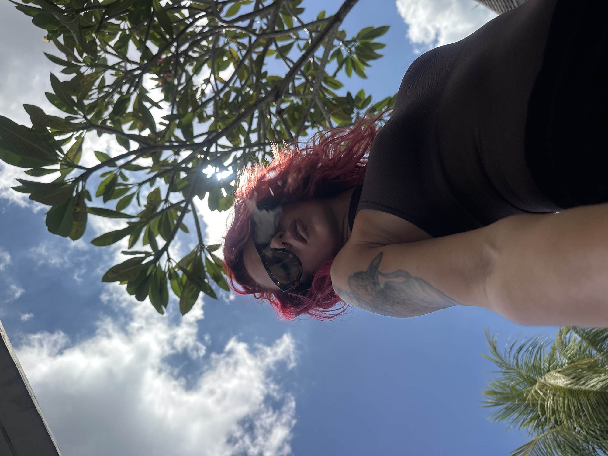 The image is an upward-angled, low-point-of-view shot in what appears to be North Kuta, Indonesia, based on the presence of palm trees, a lush green tree with broad leaves, and a partially visible building edge in the bottom left. A person with red hair, sunglasses, and a dark top is visible in the frame. The person is tilted at an angle, looking downwards, with their left arm visible and featuring a tattoo. The background includes a blue sky with scattered white clouds. The sun shines through the leaves of the tree in the top left, creating lens flare. A partial view of a palm tree is visible in the lower right corner.