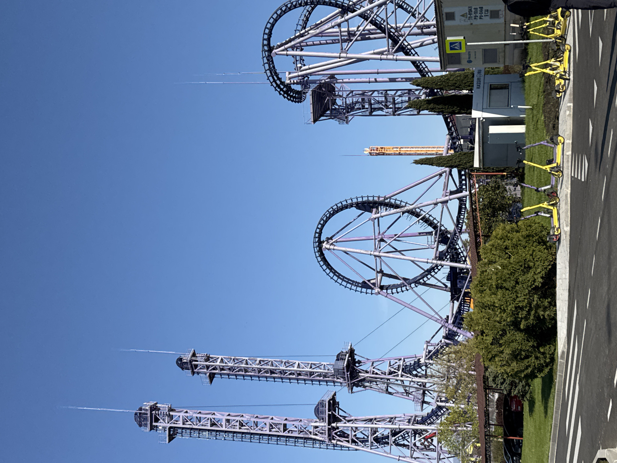 The image captures a theme park setting in Sochi, Russia, likely a recreational area. The scene is dominated by tall amusement park rides, including roller coasters with prominent loop structures and vertical towers that eject water at the top. The rides have a predominantly metal and purple construction, with a network of support beams. In the foreground, there are visible yellow rental bikes arranged along the edge of a pavement, suggesting a tourist-oriented location. There are administrative structures or kiosks. The sky is clear, and the overall environment suggests a bright, sunny day.