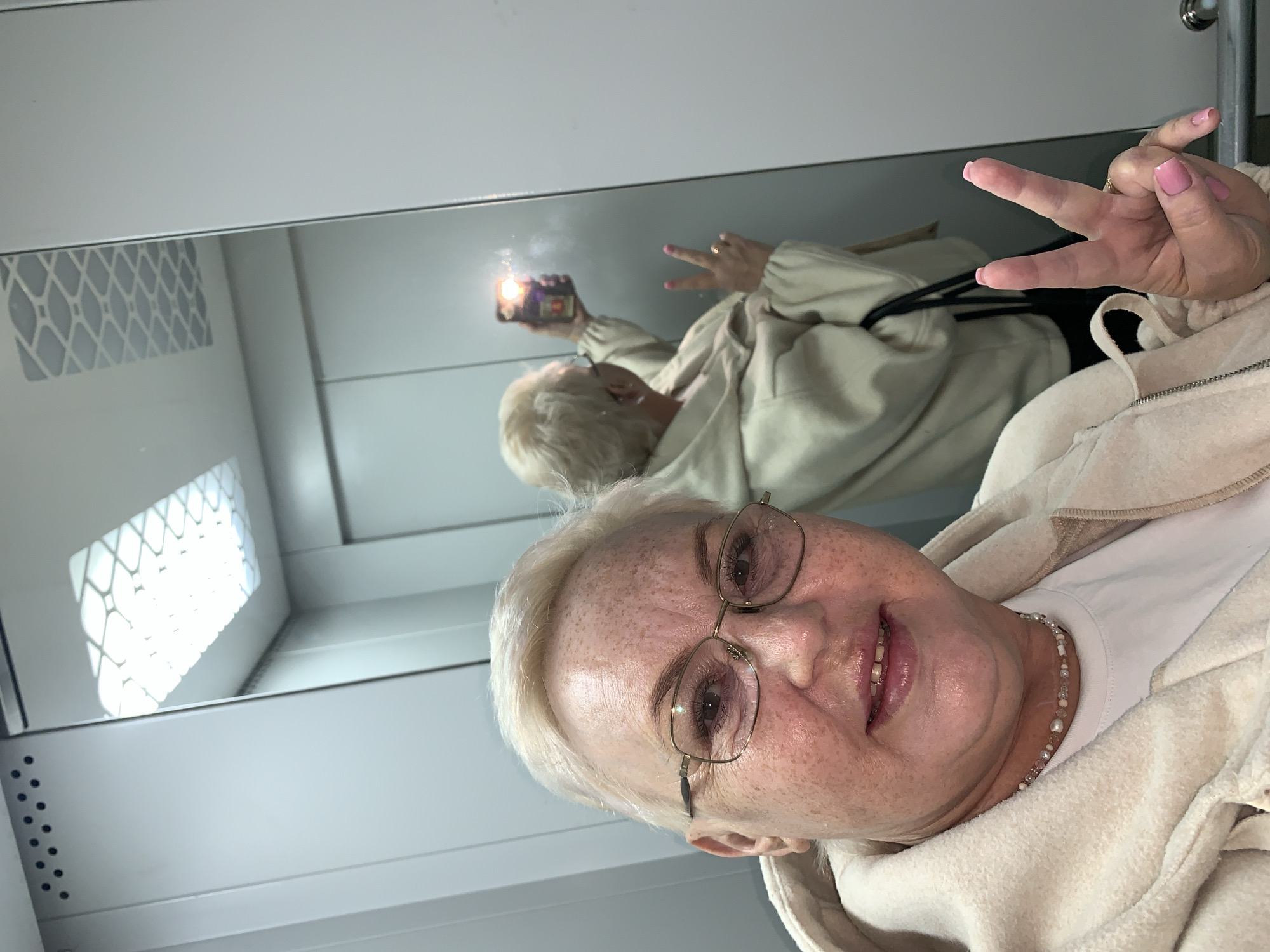 The photograph shows an older woman taking a selfie in what appears to be an elevator. She has short blonde hair, glasses, and is wearing a light-colored hoodie and a beaded necklace. She is holding up a peace sign with her right hand. Her reflection is visible in the mirrored wall behind her, where she is seen holding her phone to take the picture. The elevator has white paneled walls with some ventilation grids. The location in the city of Chaikovsky, Russia, cannot be determined from the image.