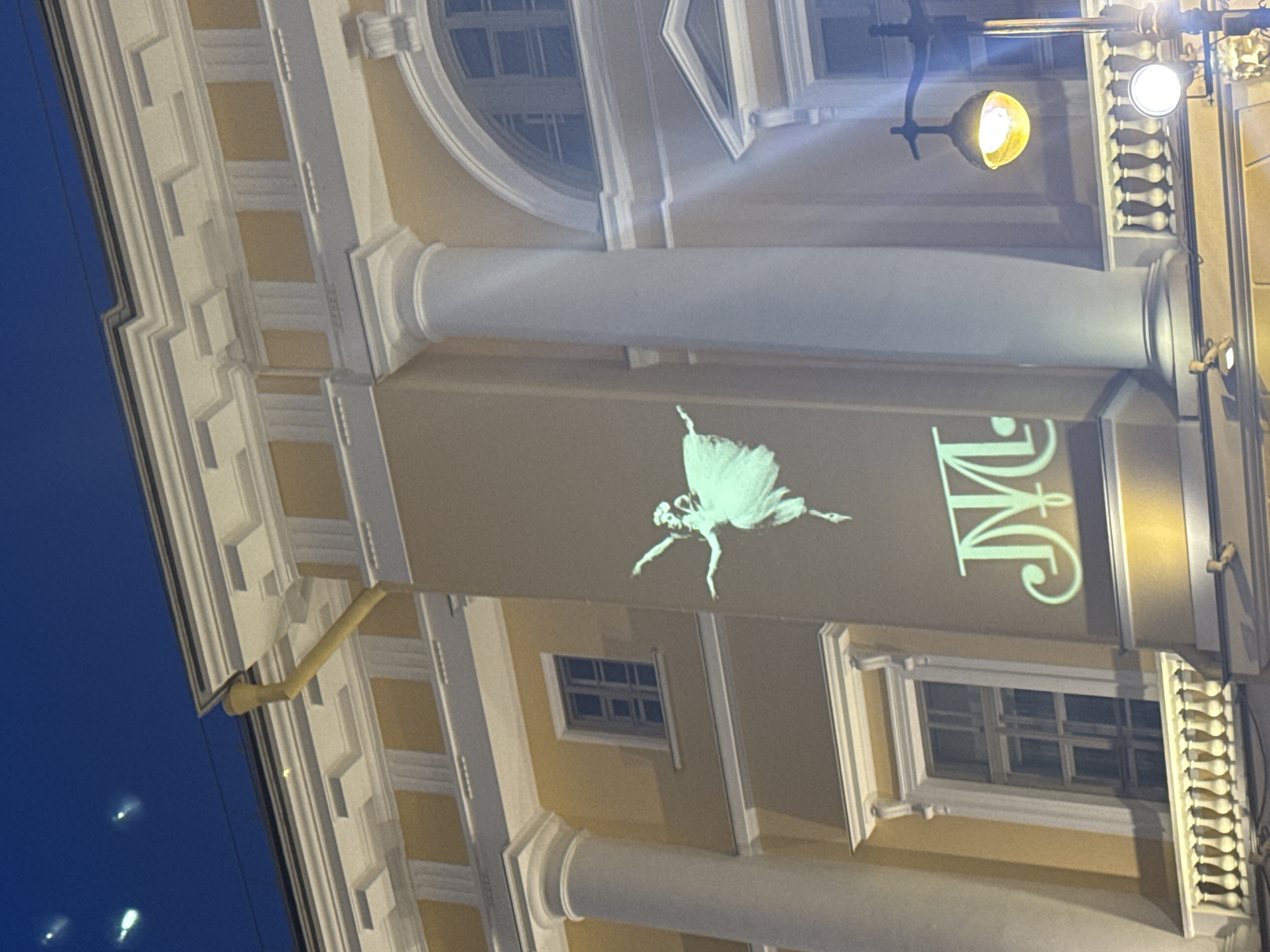 Here is a description of the image:
This is an exterior shot of a building in Saint Petersburg, Russia, displaying a ballet projection on the side. The architectural style suggests a historical building with a classical design, including a colonnade and intricate detailing. The projection consists of a ballet dancer and Cyrillic characters. The illumination around the building and the sky suggest the photo was taken at dusk or night. A street light provides further illumination.
