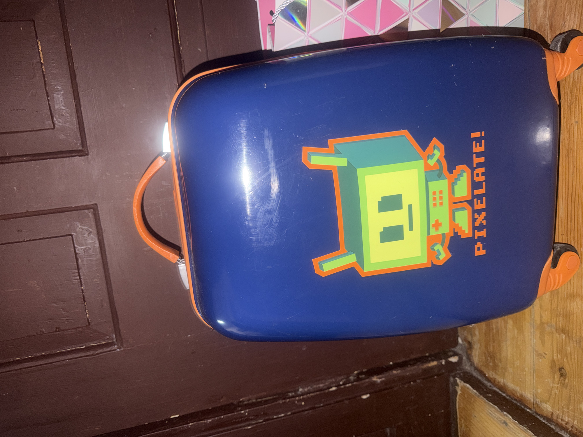 The image shows a blue suitcase with an orange trim and handle. The suitcase has a graphic design of a pixelated green monster with an orange outline and the word "PIXELATE!" in orange text. The suitcase is located on a surface next to a brown paneled door and what appears to be unfinished wood. A portion of a bag with a pink and white geometric design is visible in the upper left corner.