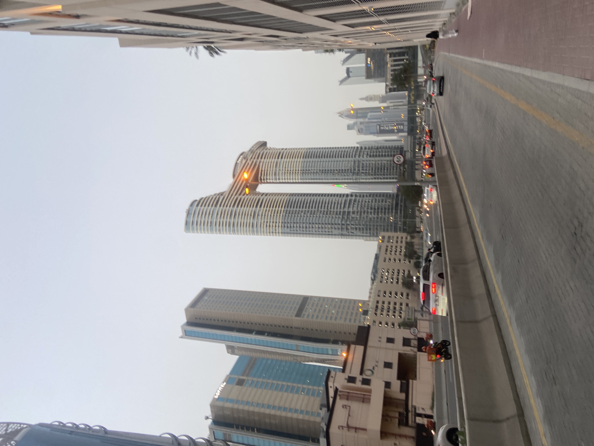 The image shows a street scene in Dubai, United Arab Emirates, featuring distinctive modern architecture. Prominently visible are two cylindrical towers connected at the top. Below those is another building with a sign reading "SOFITEL". The image is taken from street level, with a roadway and sidewalk visible on the right. There is moderate traffic on the road, with various cars and a motorcycle with an orange box on it. The sky is overcast. Other buildings are visible in the background, giving the impression of a dense urban environment. There is a speed limit sign visible on one of the towers with the number 60.