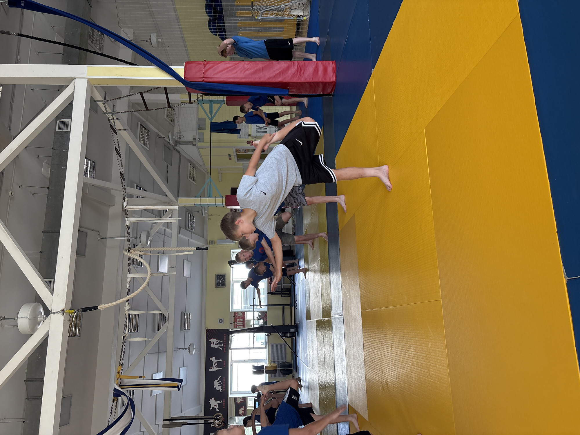 In a sports hall, likely in Chaikovsky, Russia, several young males are engaged in a physical training session on a blue and yellow mat. The lighting is bright, indicating an indoor environment with fluorescent ceiling lights visible. The walls are yellow, and various sports equipment and training aids are present, including what appears to be a rowing machine, weight racks, and ropes suspended from the ceiling. Several boys are in mid-movement, some stretching or performing balancing exercises, while others observe. A red padded post is also visible. The overall atmosphere suggests a martial arts or athletic training facility.