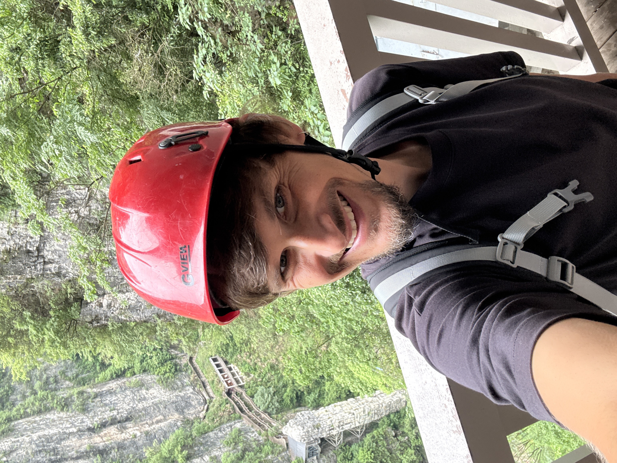 A man wearing a red helmet and a dark shirt with gray straps is smiling at the camera. He appears to be standing on a structure with white railings. In the background, there is a lush green landscape with rocky formations and trees. A bridge or platform structure is visible in the distance. The location is likely in Zhangjiajie, China, given the unique geological formations in the area.